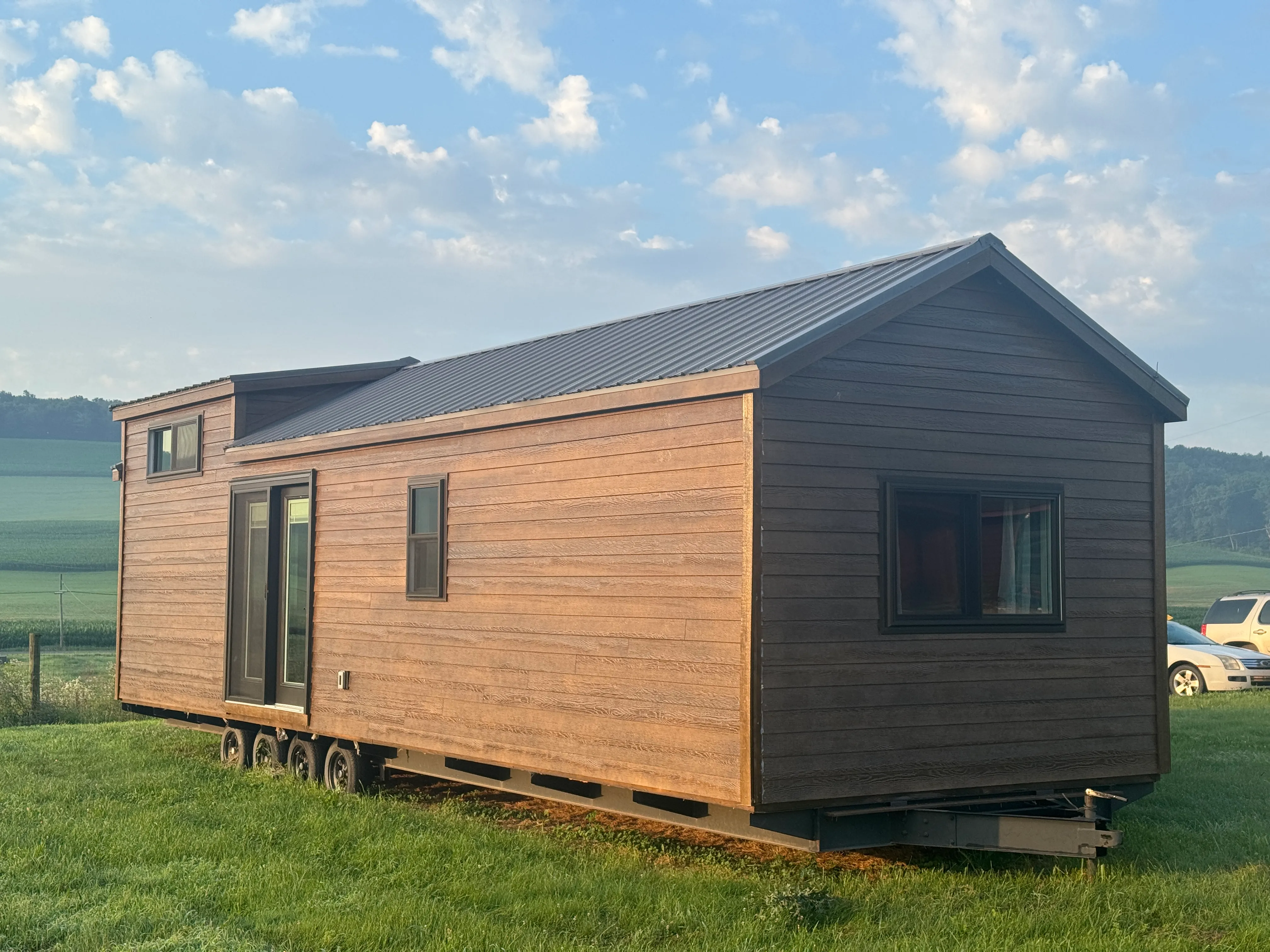 A wooden tiny house sits on grass with a field and hills in the background under a partly cloudy sky.