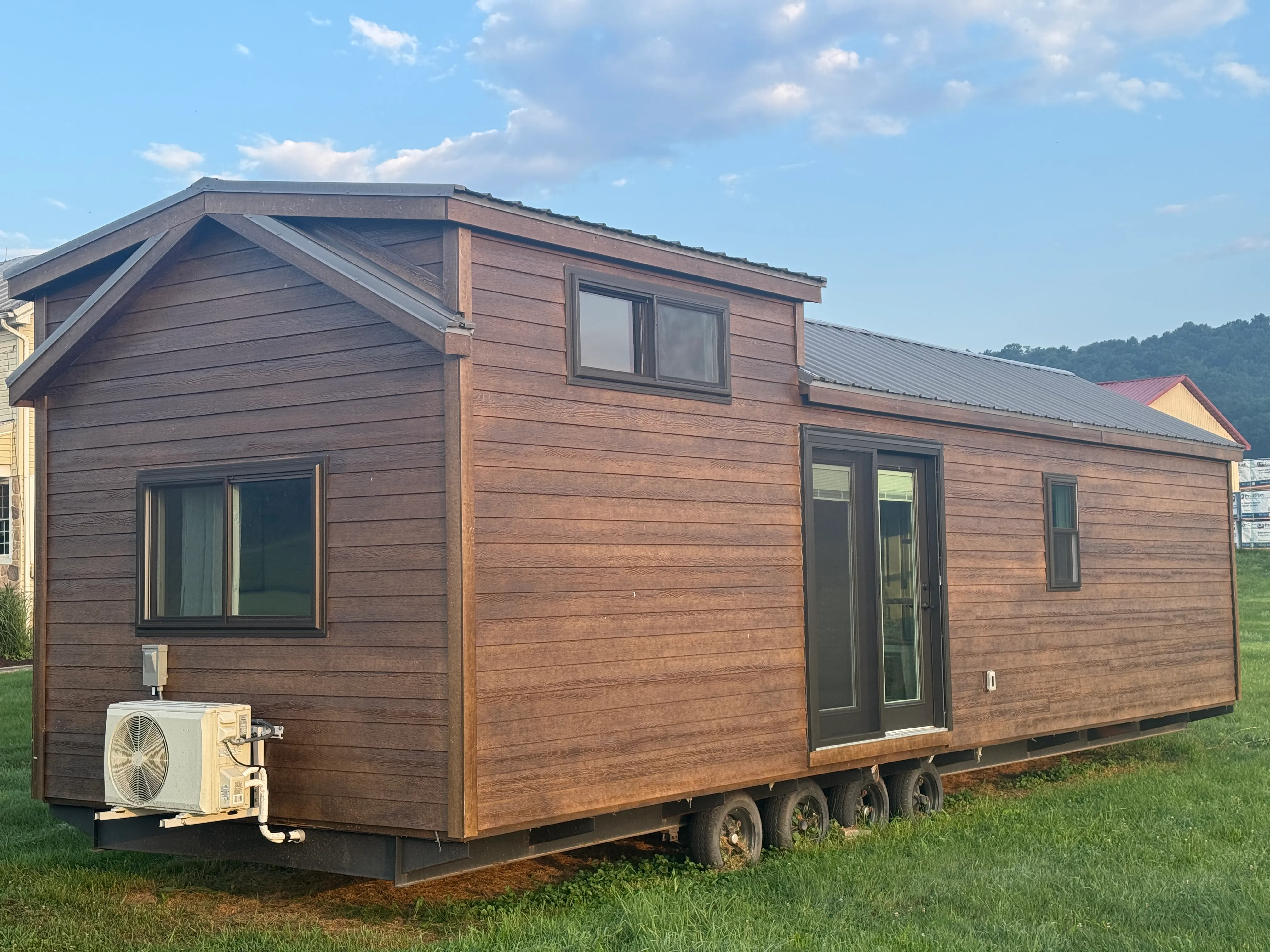 A brown tiny house sits on grass, featuring multiple windows, a glass door, and an external air conditioning unit.