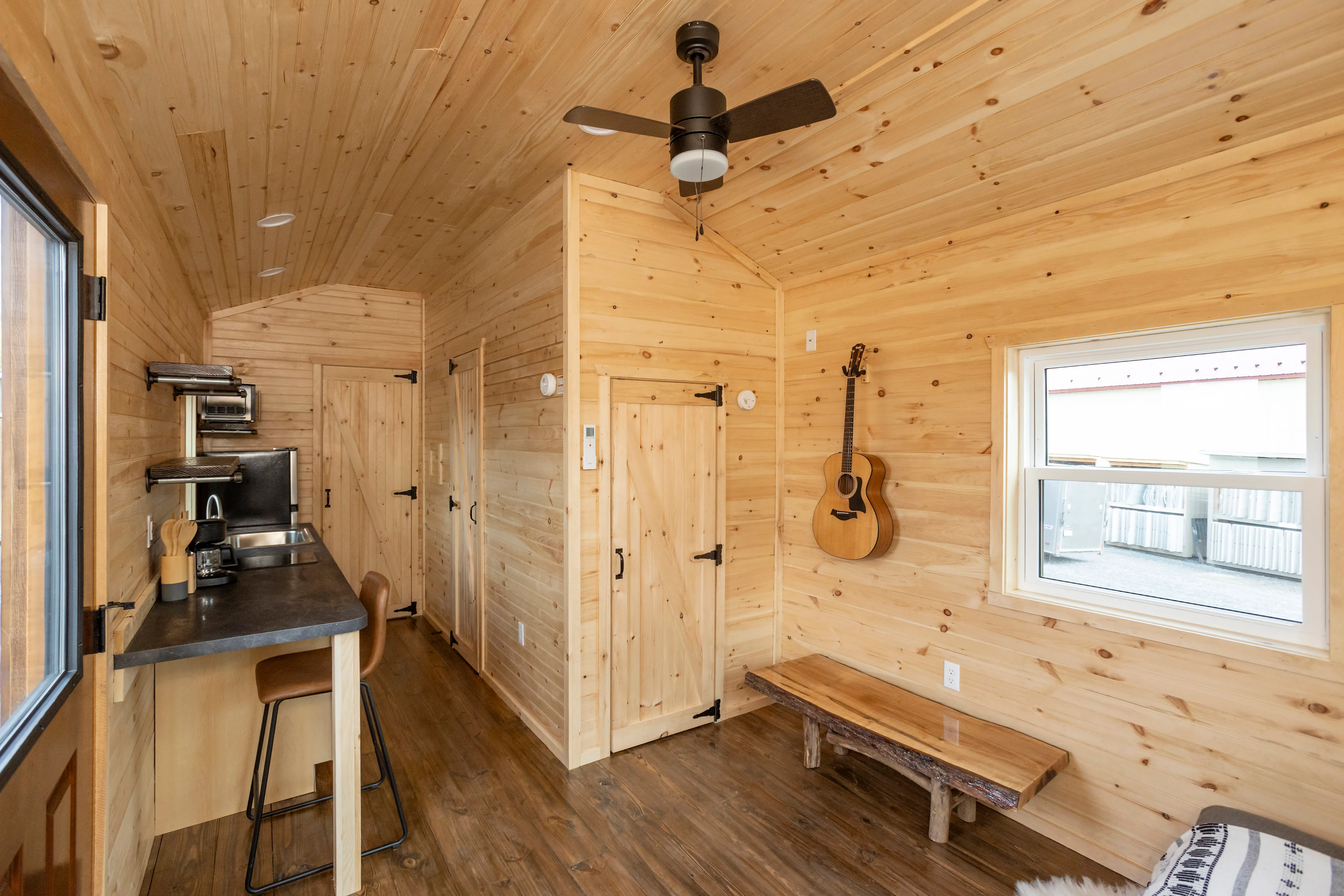 Interior of a small wooden cabin with a kitchenette, bar stools, ceiling fan, guitar on the wall, bench, and window. Light wood paneling covers the walls and ceiling.
