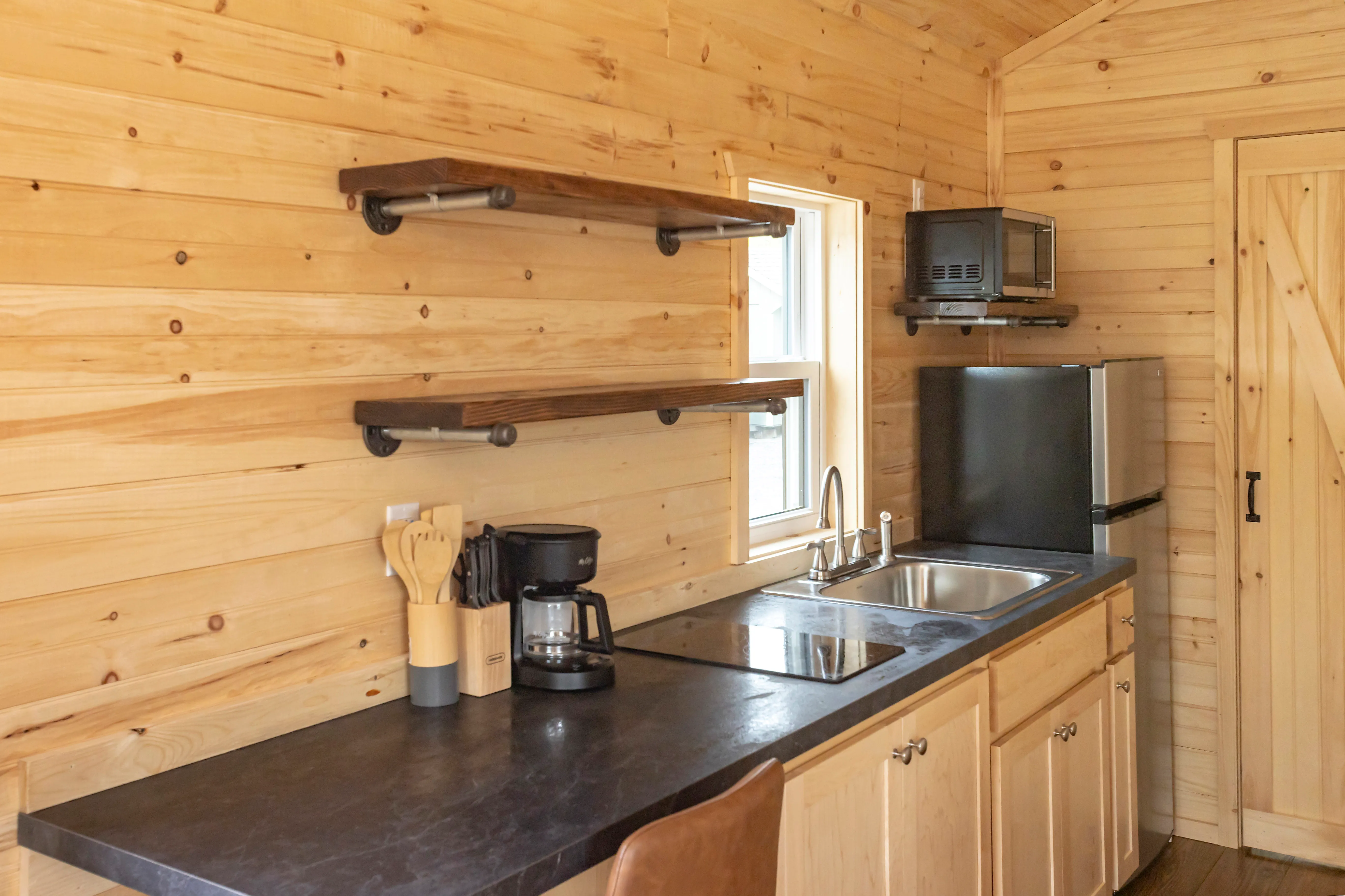 Small kitchen with light wood walls, open shelves, a sink, black countertop, coffee maker, mini fridge, microwave and window above the sink.