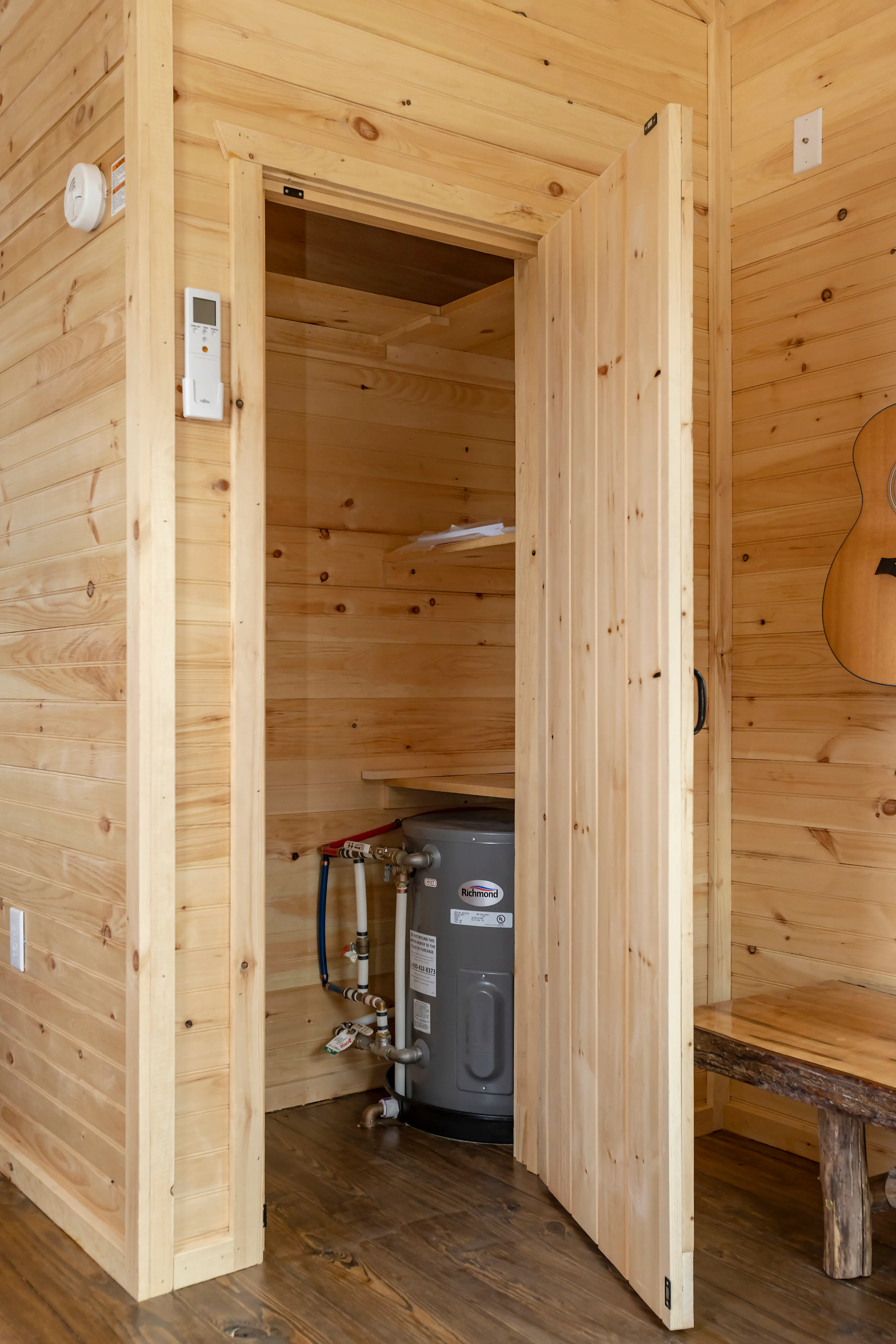 A small closet with an open wooden door reveals a water heater inside a room with wooden walls and floors.