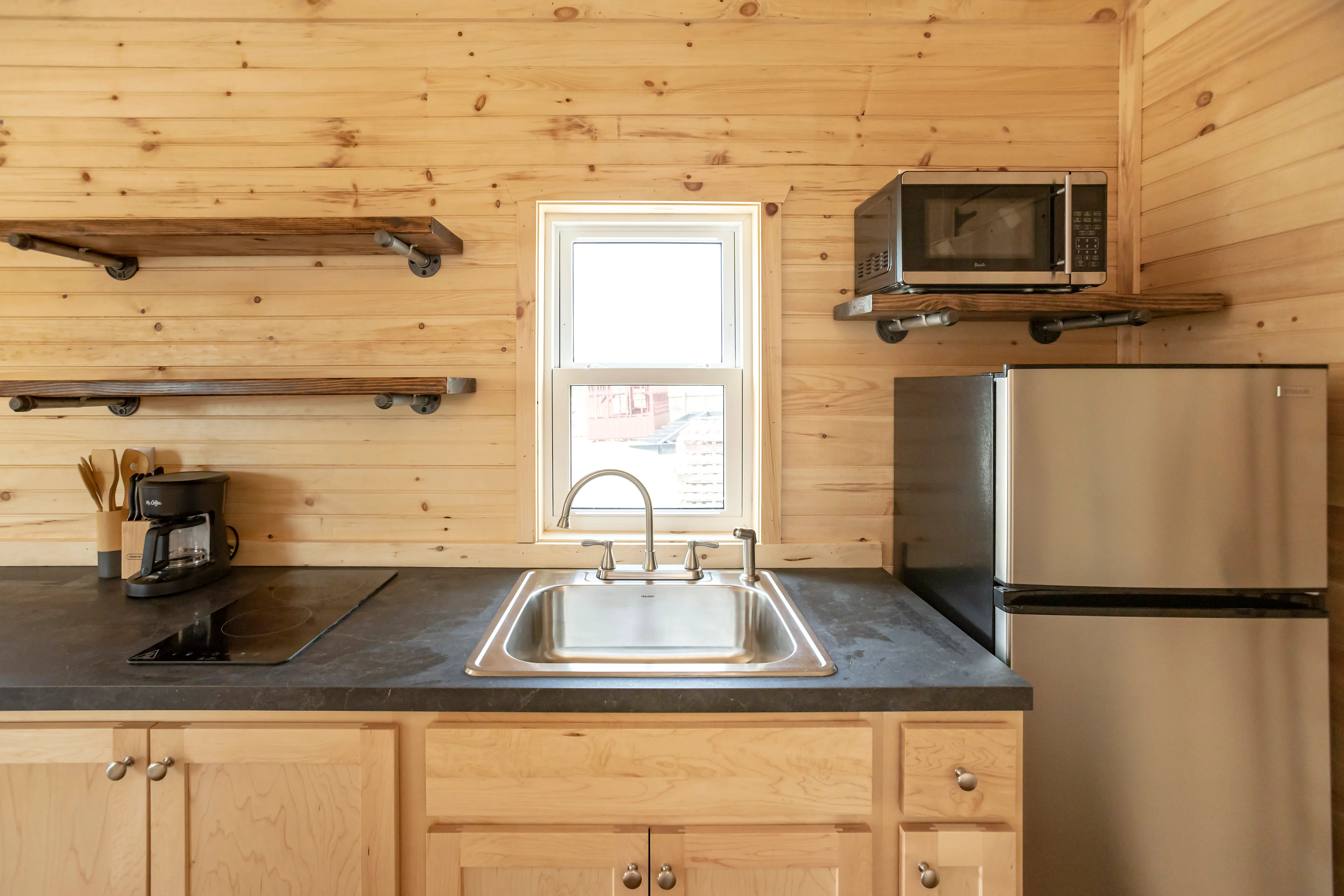 Compact kitchen with light wood cabinets, a stainless steel sink, small refrigerator, microwave, coffee maker, electric cooktop, and open shelving on a wood-paneled wall.