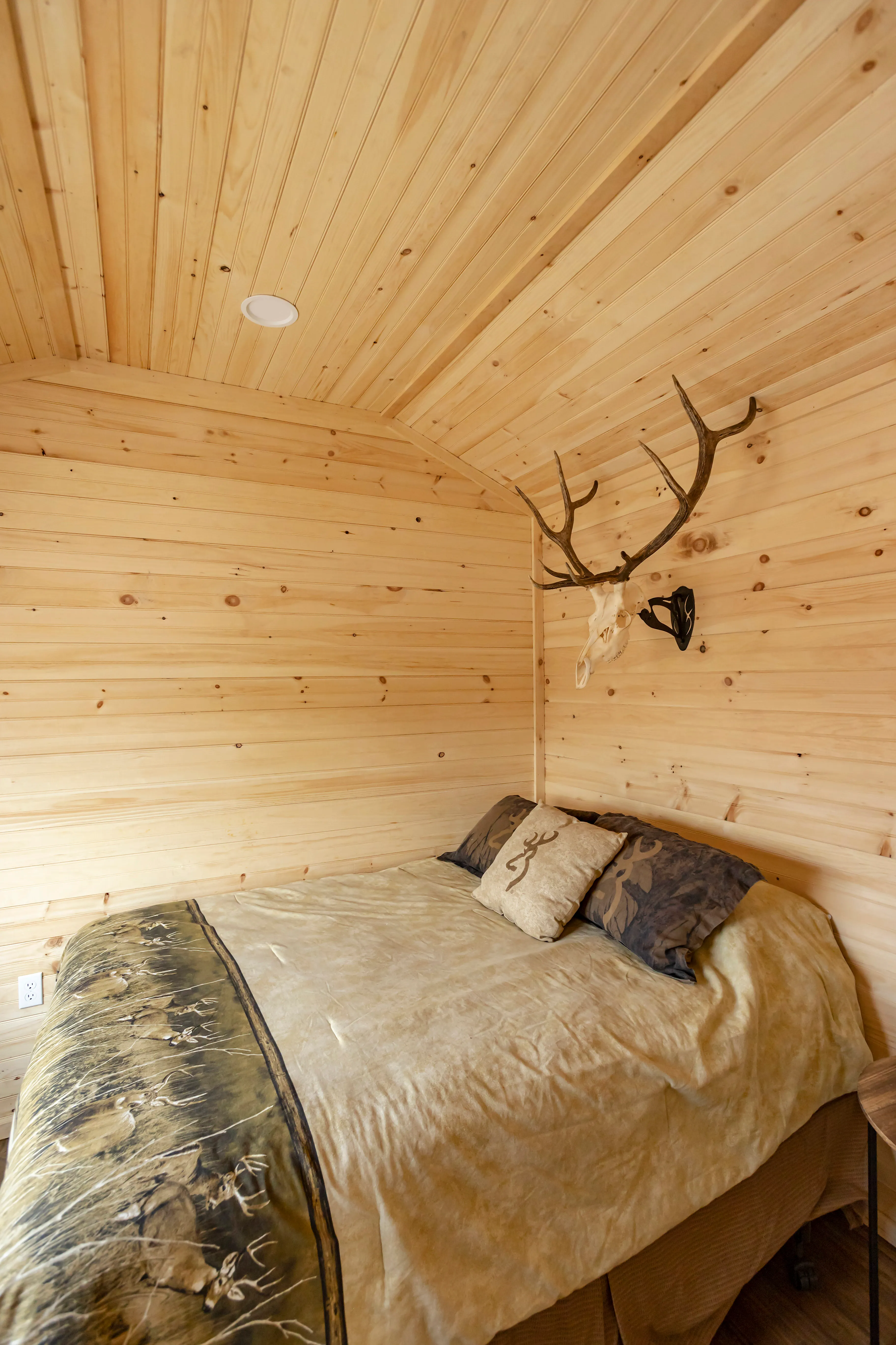 A small bedroom with wood-paneled walls, a bed with decorative pillows and blanket, and a mounted antler decoration above the headboard.