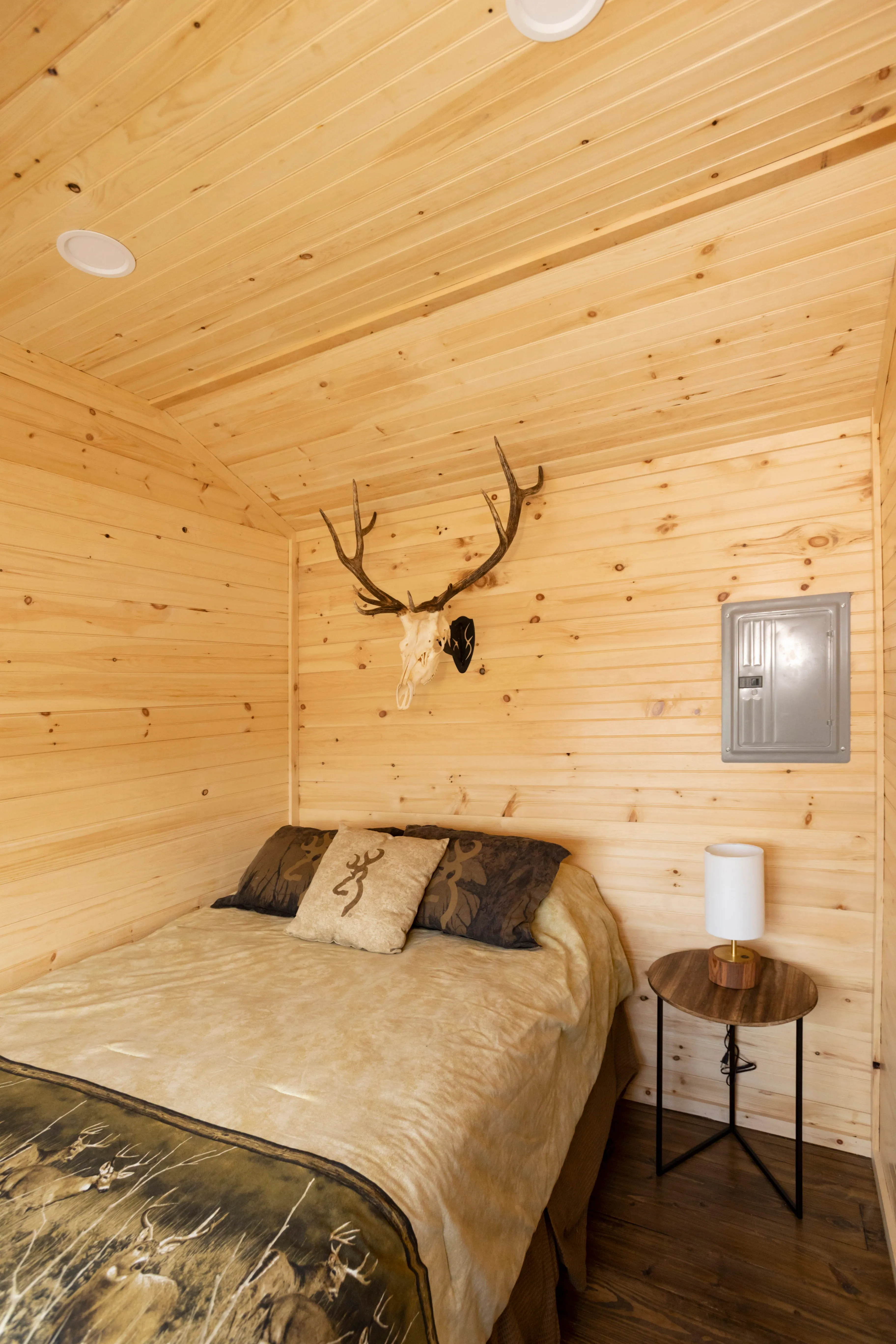 Small bedroom with wood-paneled walls and ceiling, a bed with animal-themed bedding, a round side table with a lamp, and a deer skull with antlers mounted above the bed.