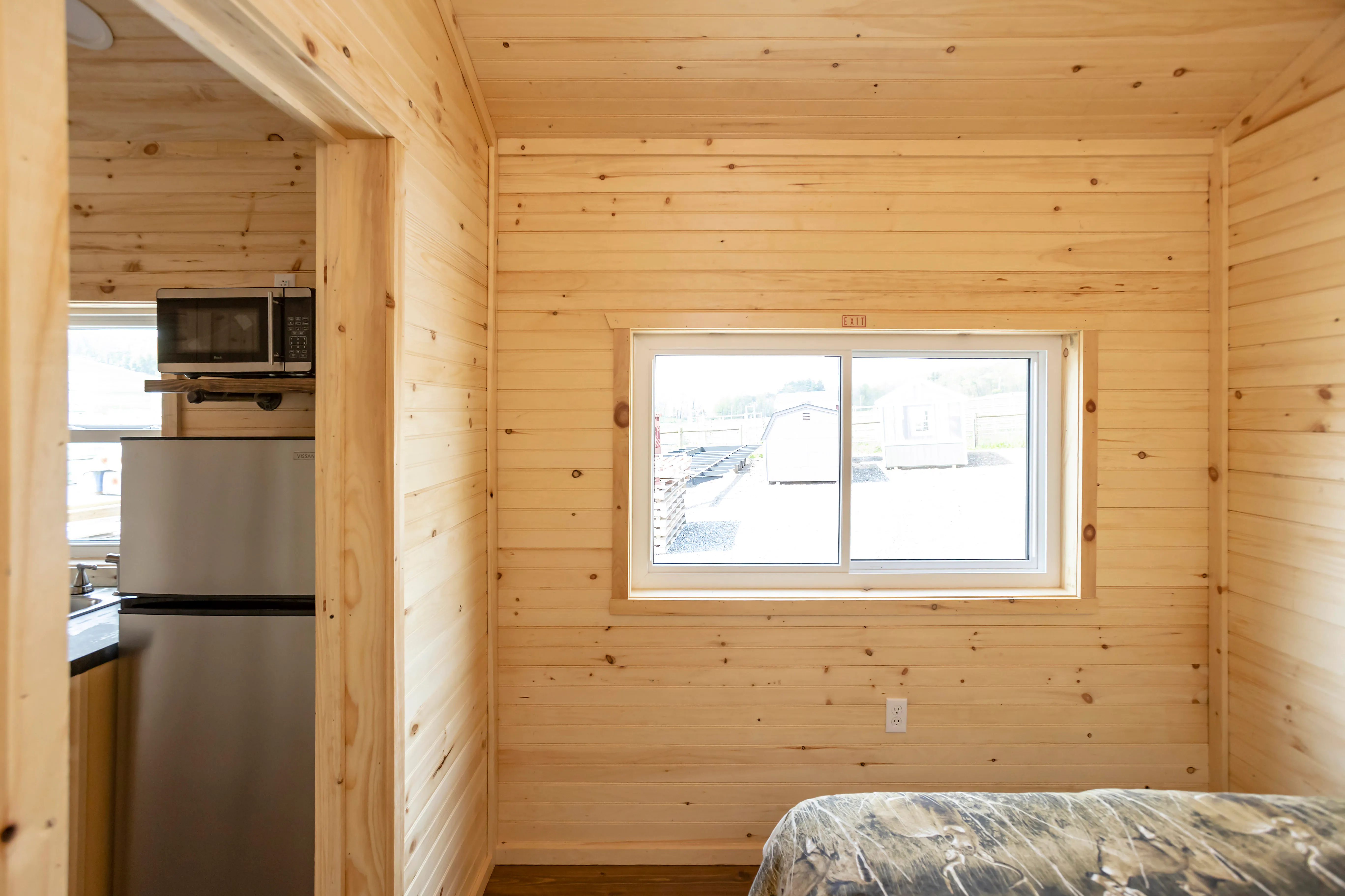 Small room with light wood paneling, a window, and part of a bed visible in the lower right corner. A refrigerator and microwave are seen in the adjoining area.