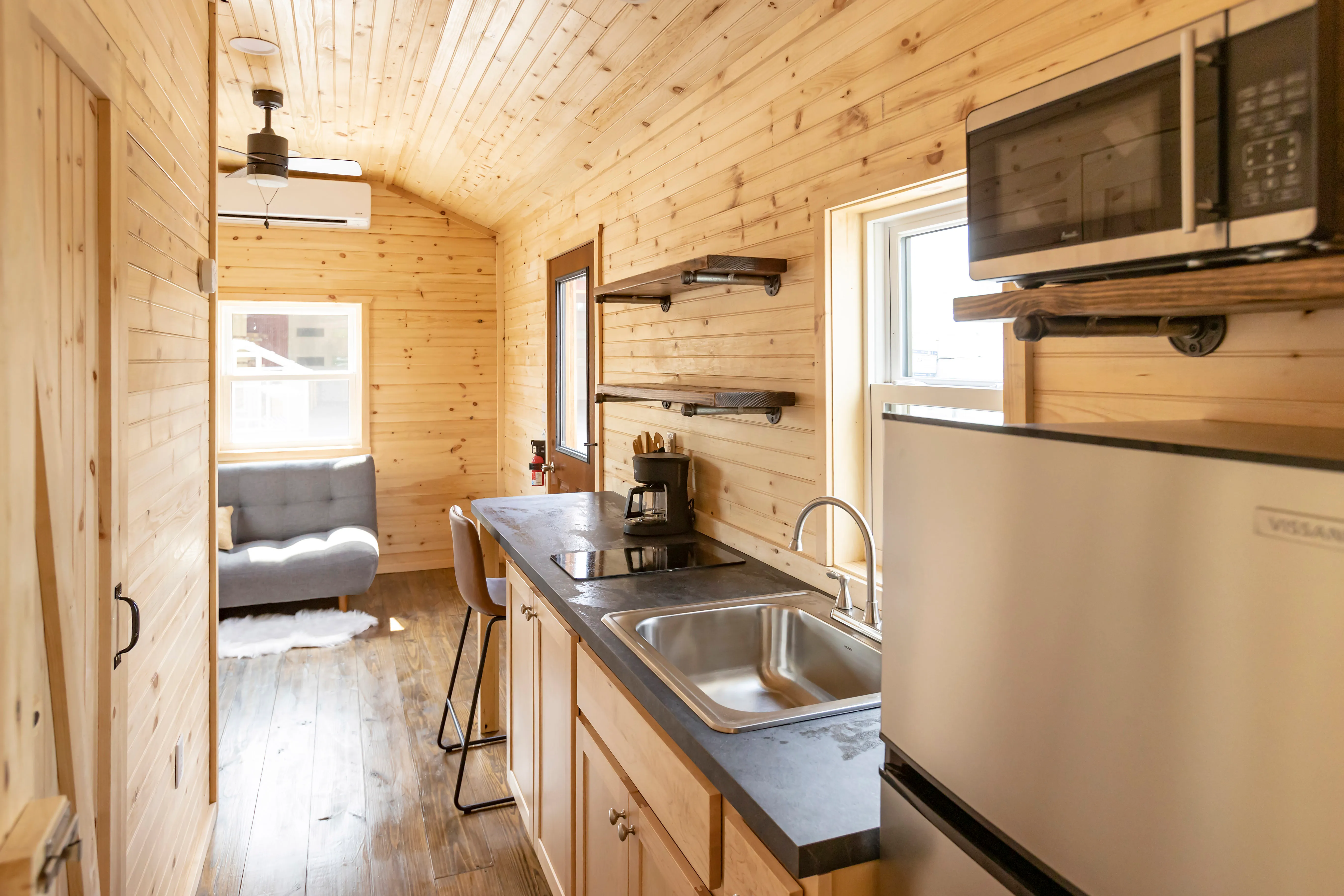 Interior of a small wooden cabin kitchen with a stainless steel sink, fridge, microwave, open shelves, a coffee maker, and a sitting area with a gray sofa at the far end.