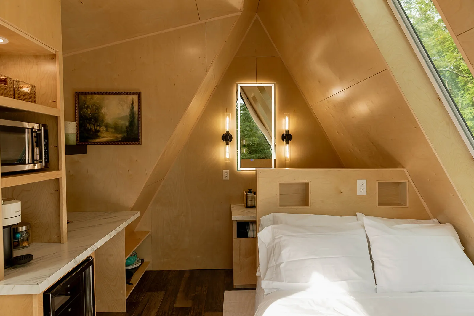 A modern A-frame cabin interior with wood-paneled walls, a neatly made bed, built-in shelves, a kitchenette, and a window.