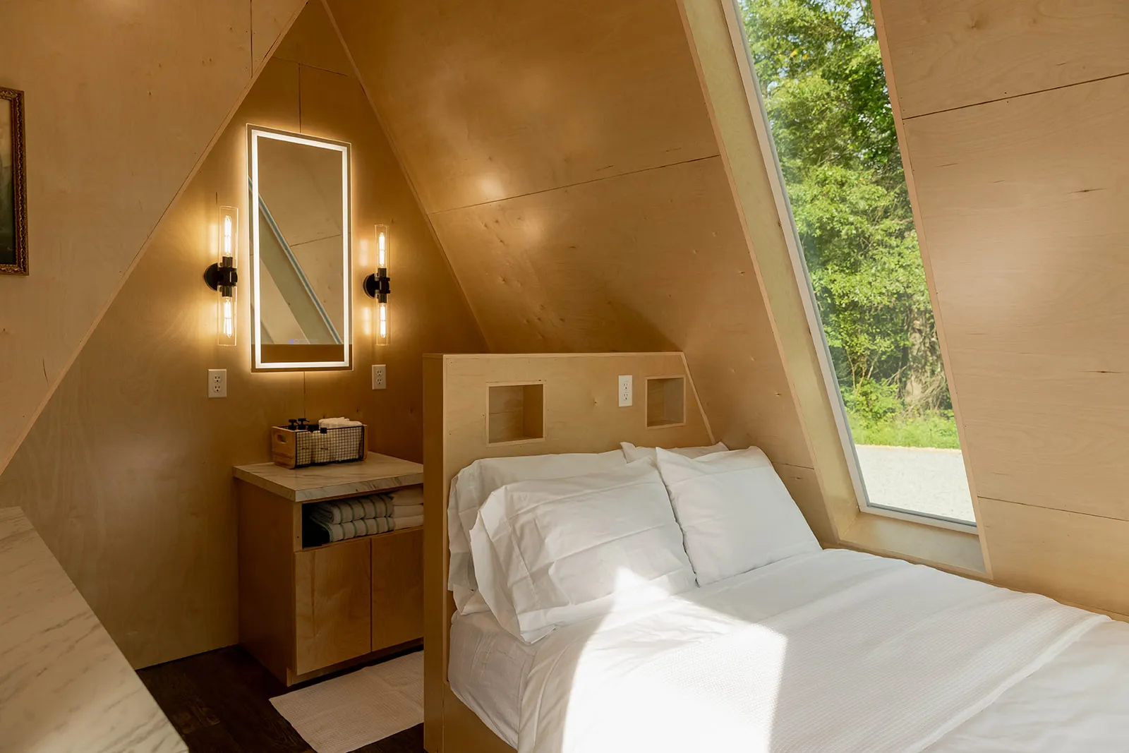 A modern A-frame cabin bedroom with wood-paneled walls, a neatly made bed, a large window, and a small vanity with an illuminated mirror.