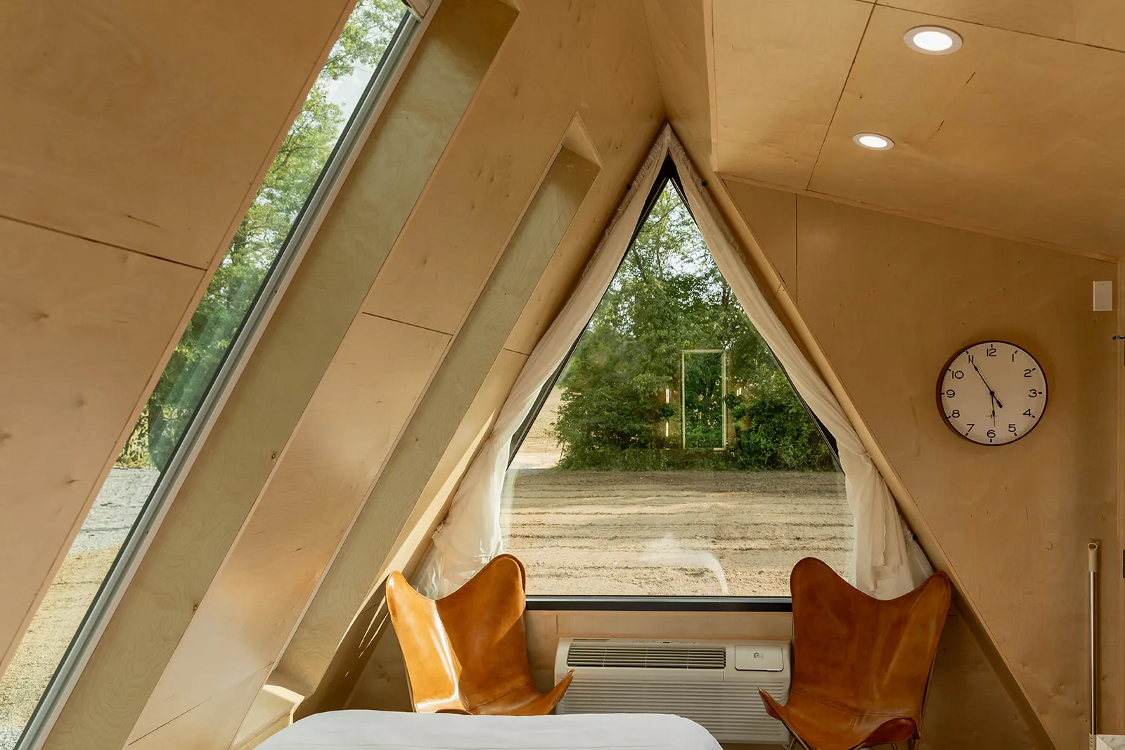 Interior of an A-frame cabin with large triangular windows, two brown leather chairs, and clock on the wall.