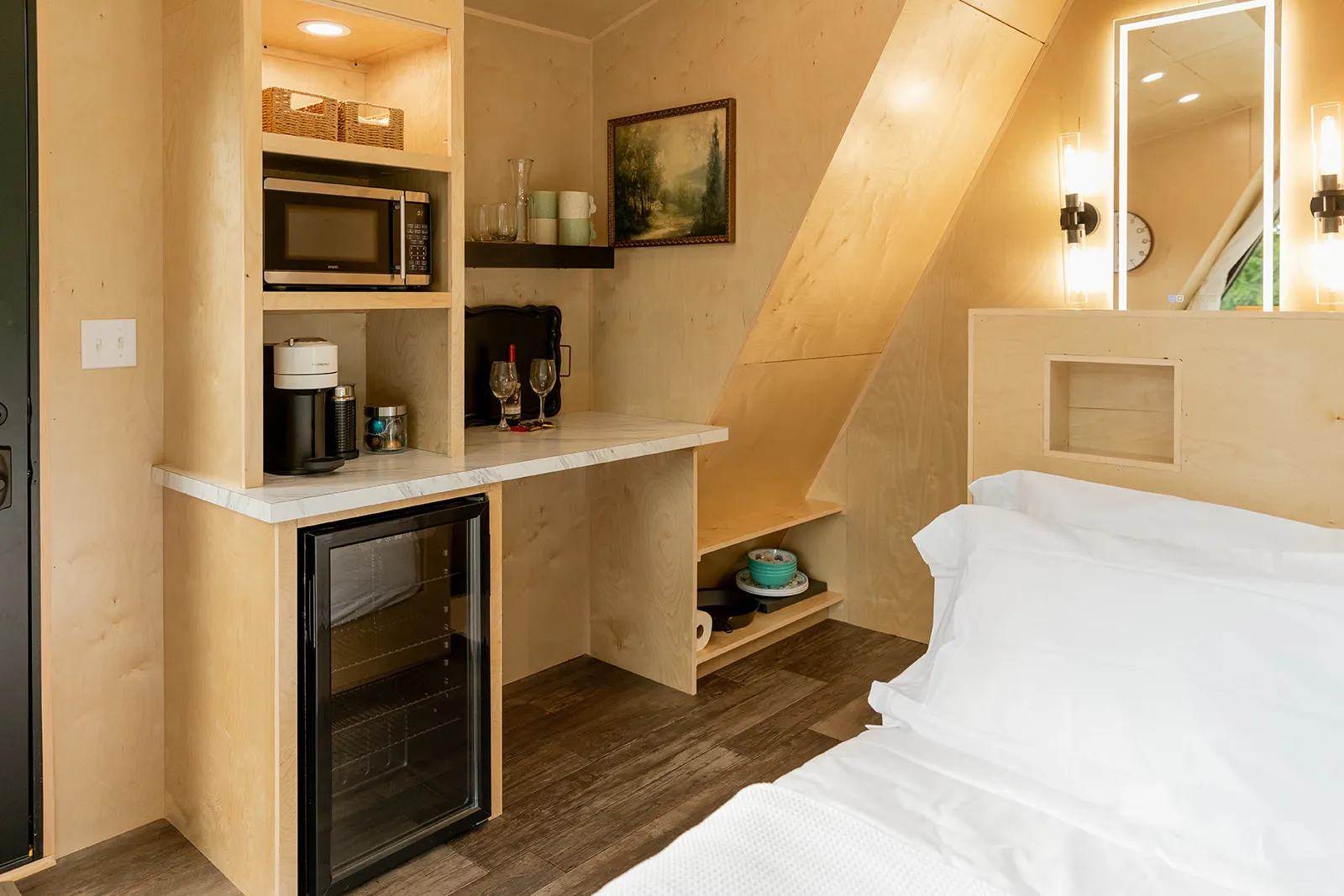Compact kitchenette in a wooden cabin with a mini fridge, microwave, coffee maker, shelves with dishes, and a made bed in the foreground.