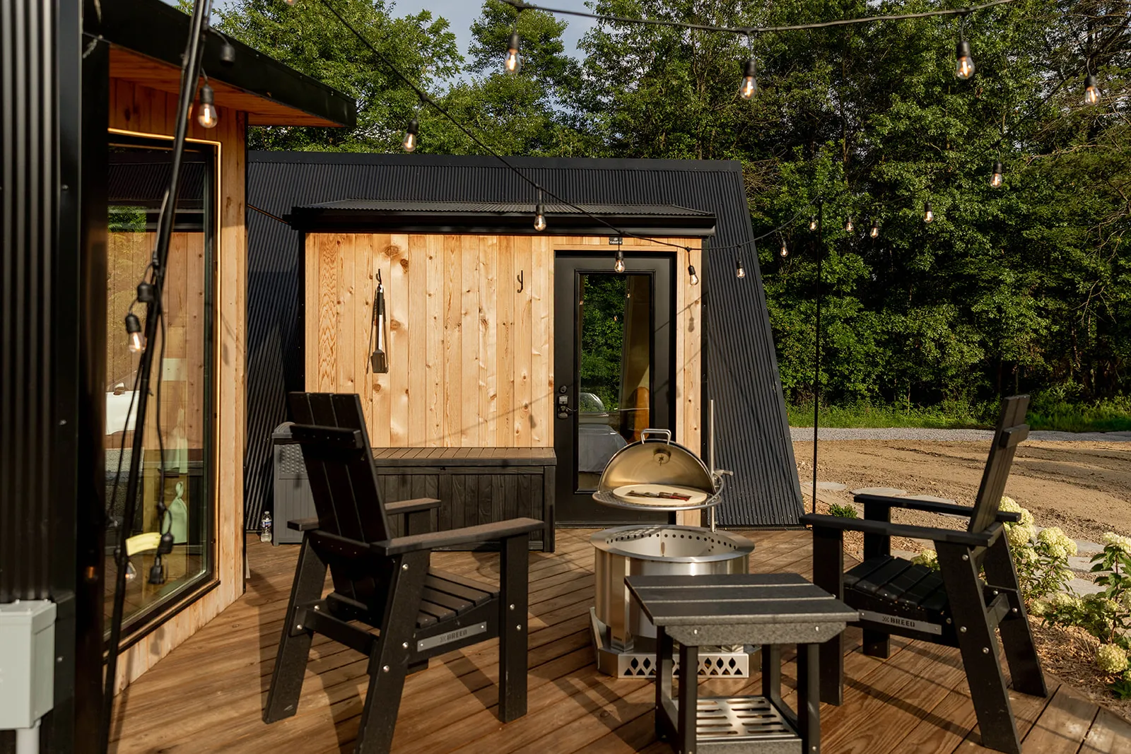 A wooden deck with four black chairs, a fire pit, and string lights, outside a modern wood-and-metal cabin.