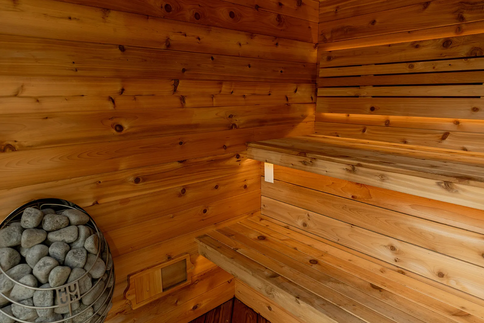 Interior of a wooden sauna with tiered benches and a heater filled with stones in the corner.