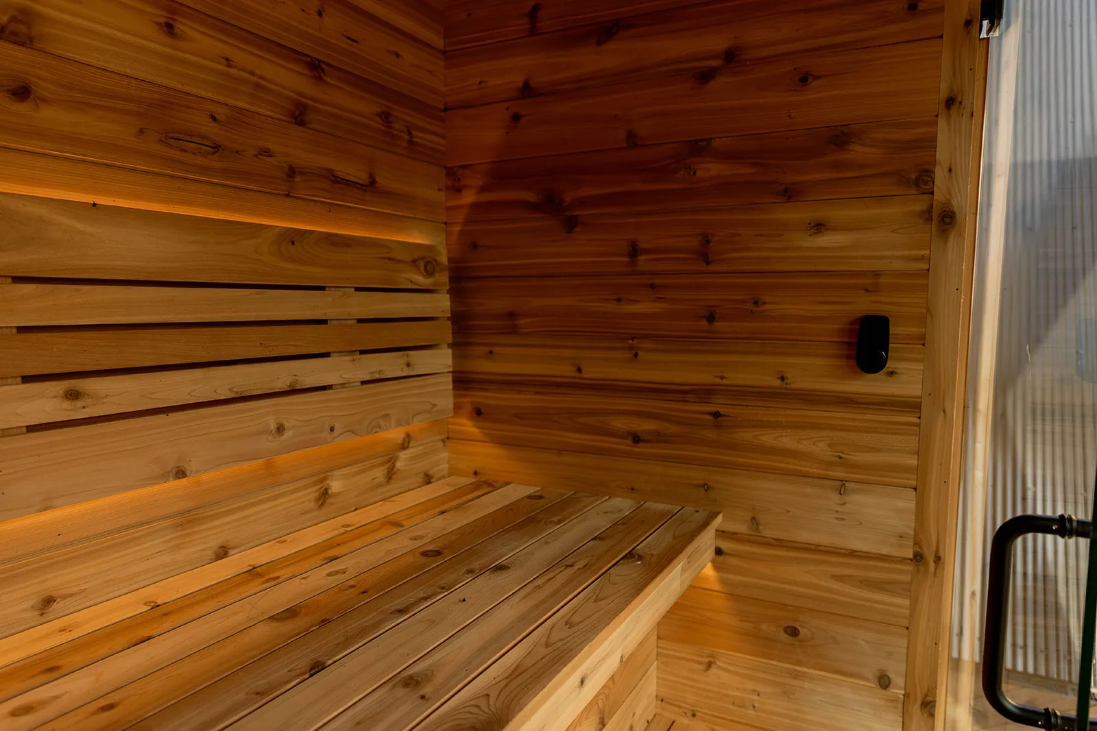Interior of a wooden sauna with a bench, warmly lit walls, and a glass door visible on the right side.