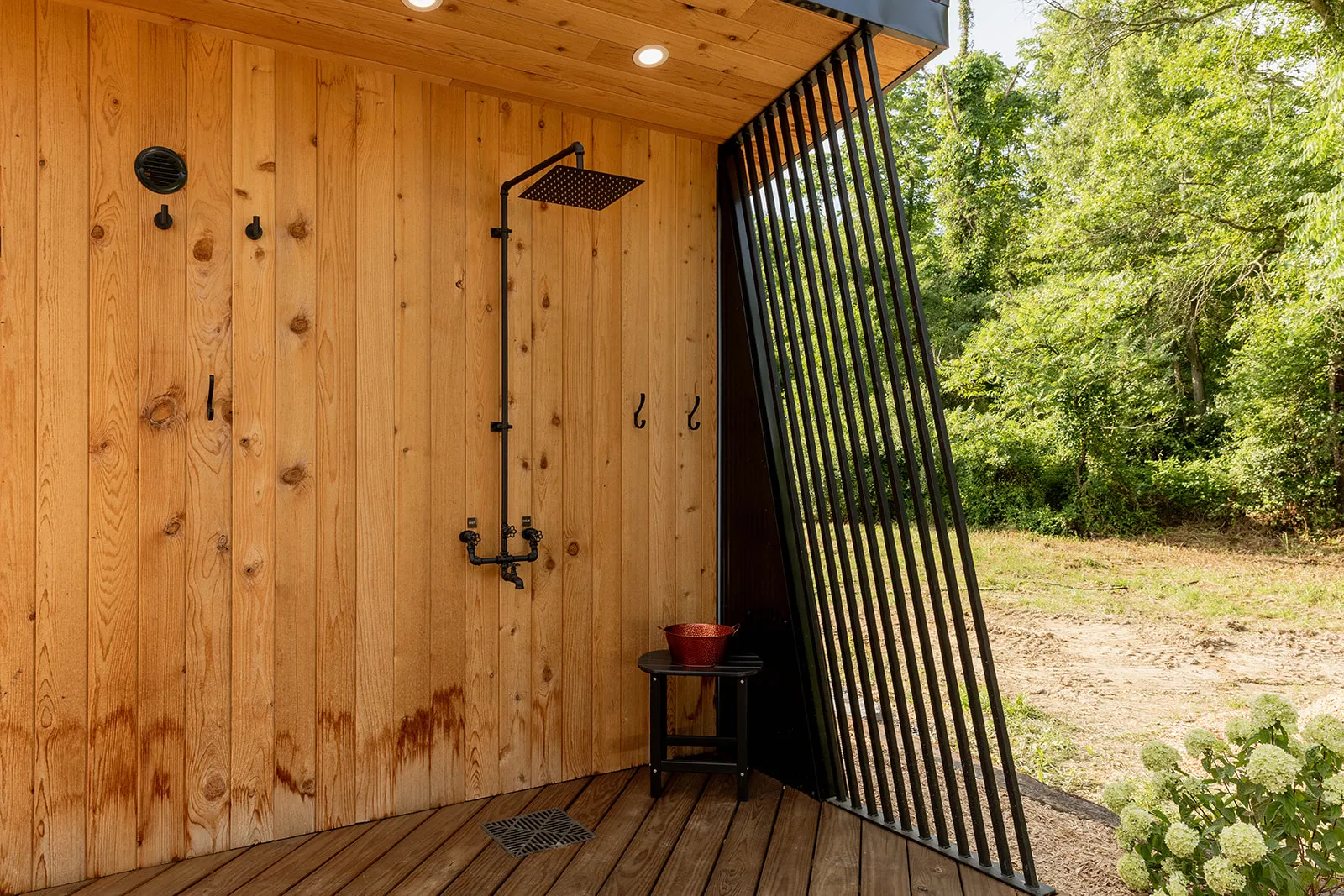Outdoor wooden shower area with a black metal rain showerhead, two wall hooks, a small black stool with a red bowl, and vertical black metal slats.