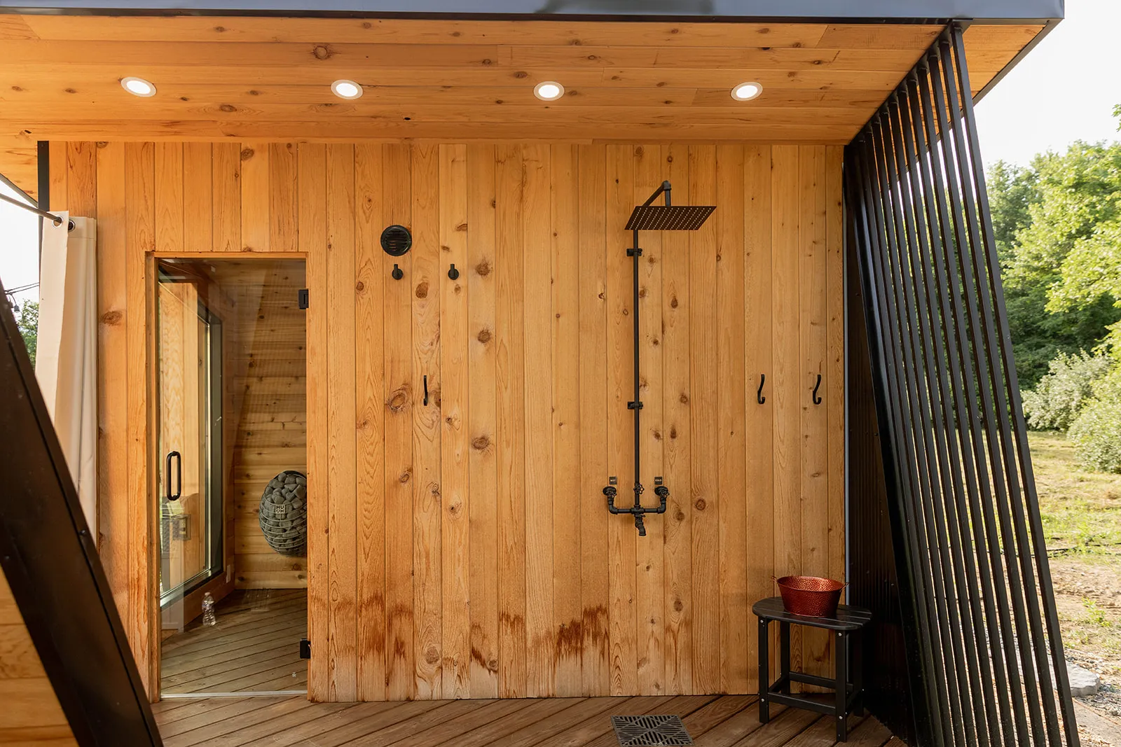 A wooden outdoor shower area with a rain shower head, metal hooks, a small stool with a red bucket, and a partially open glass door leading to sauna.