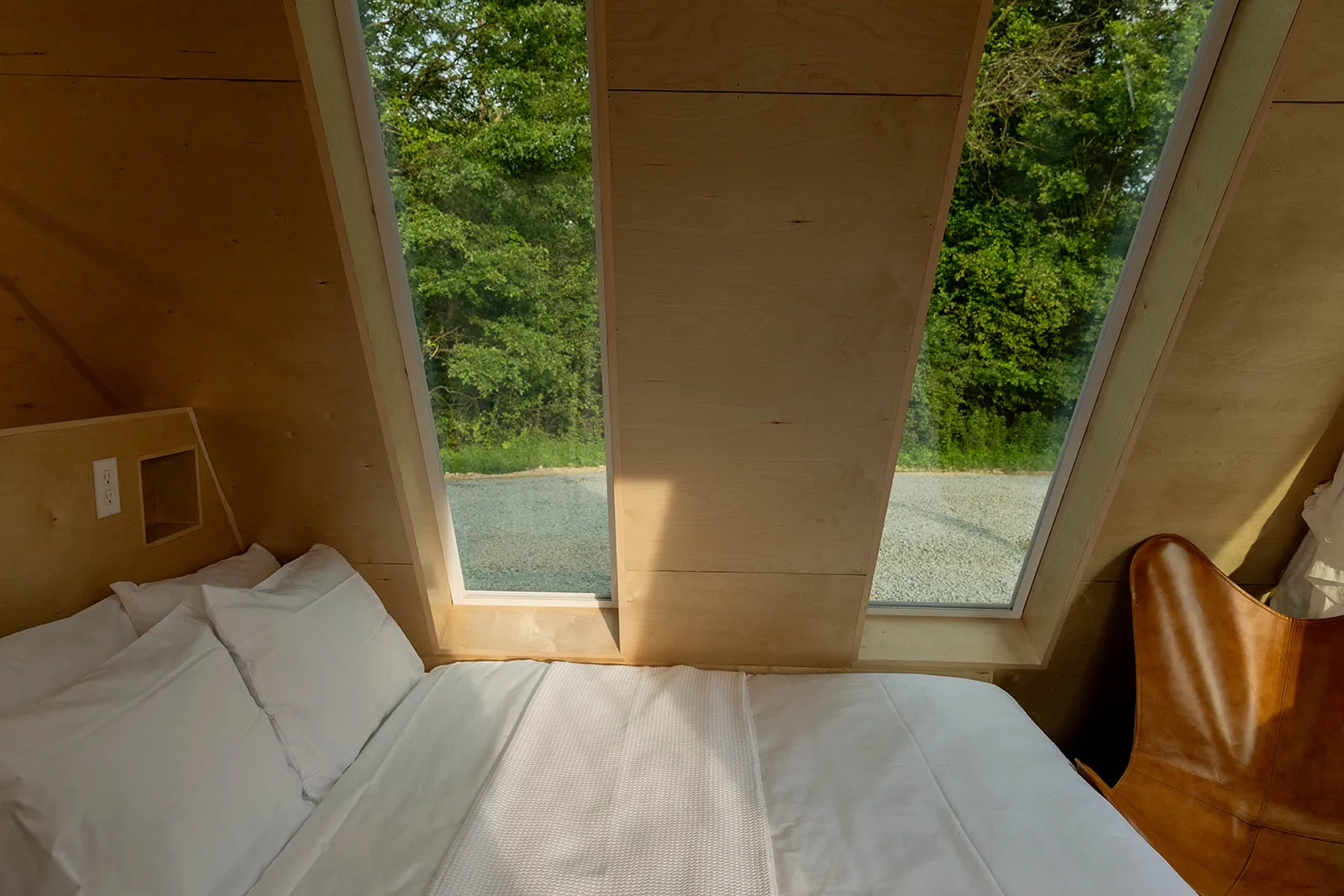 A bed with white sheets is positioned beside two large angled windows.