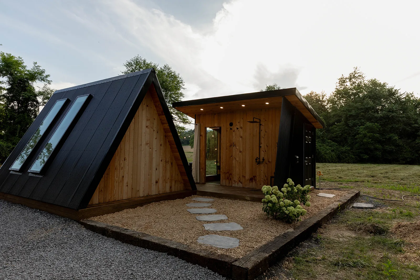 A modern A-frame cabin with wood siding, large windows, and an adjacent small structure, set on a gravel and mulched lot surrounded by grass and trees.