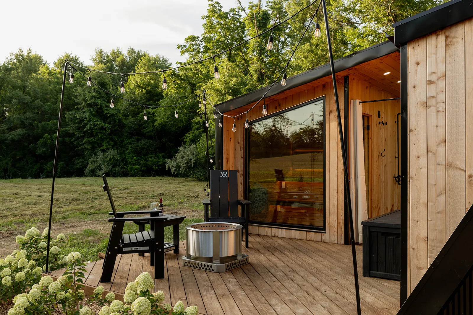 A modern wooden tiny house with a large window sits on a deck with chairs, a fire pit and string lights.