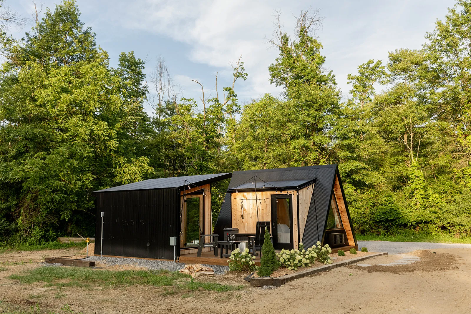 A modern black A-frame cabin with large windows and a matching building sits on a cleared plot surrounded by green trees. Outdoor seating and decorative lights are visible on the deck.