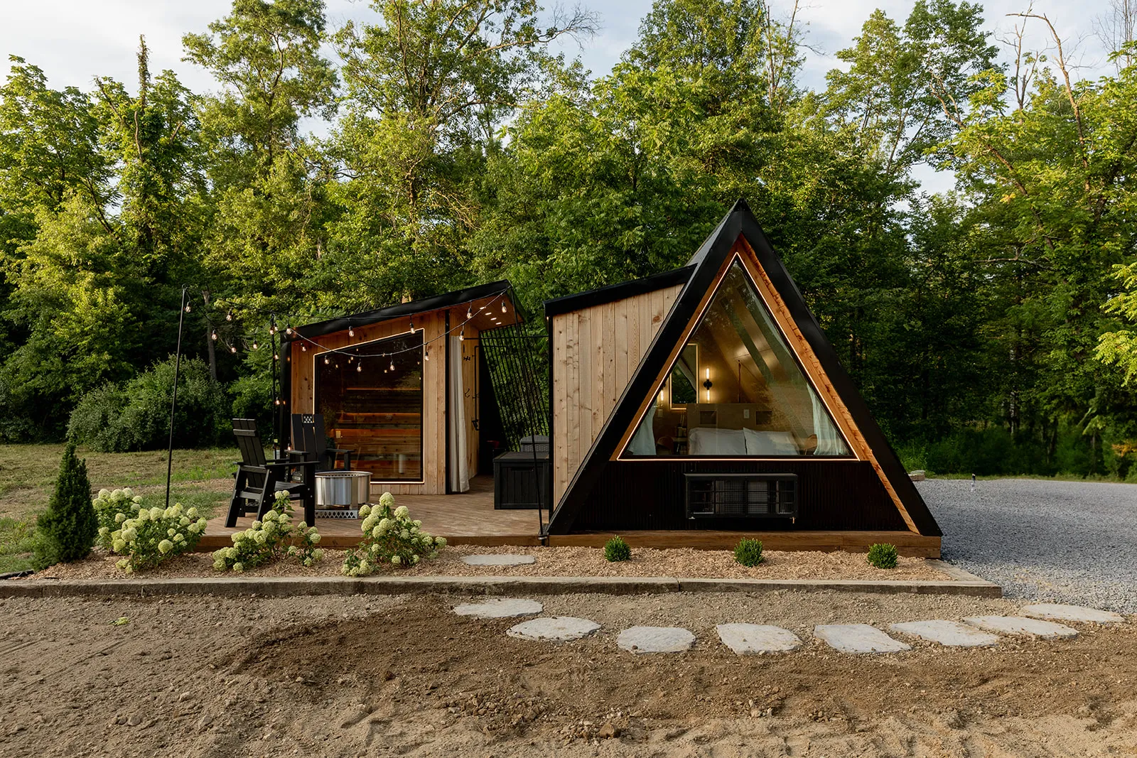 A modern A-frame tiny house with large glass window and a matching building and a outdoor seating area, surrounded by trees and a gravel path.