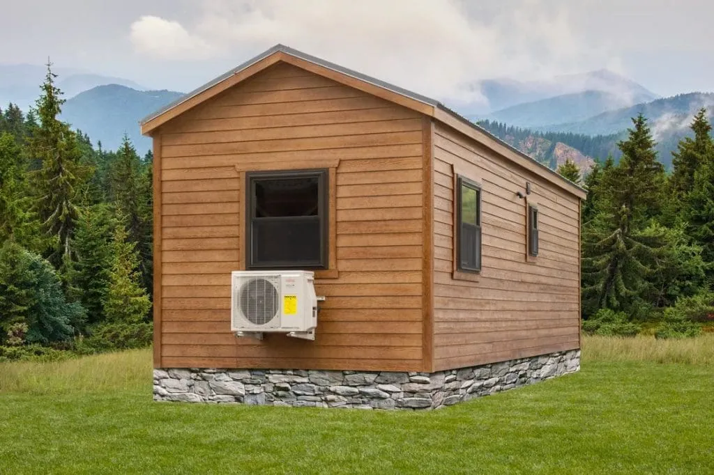 A small wooden cabin with a stone foundation, window-mounted air conditioning unit, and surrounded by grass and trees with mountains in the background.