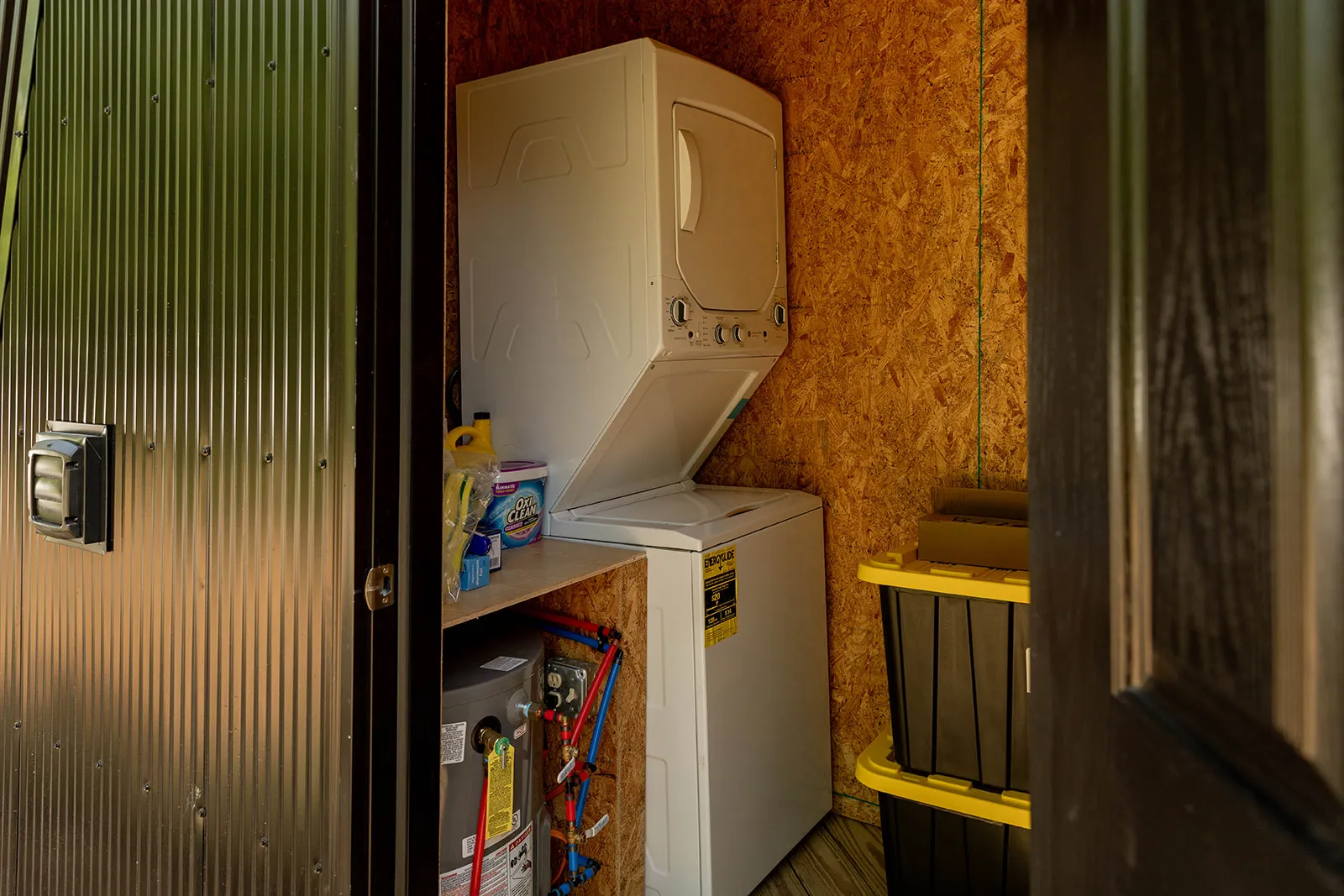 A small laundry room with a stacked washer and dryer, cleaning supplies, a water heater, and plastic storage bins against wood-paneled walls.