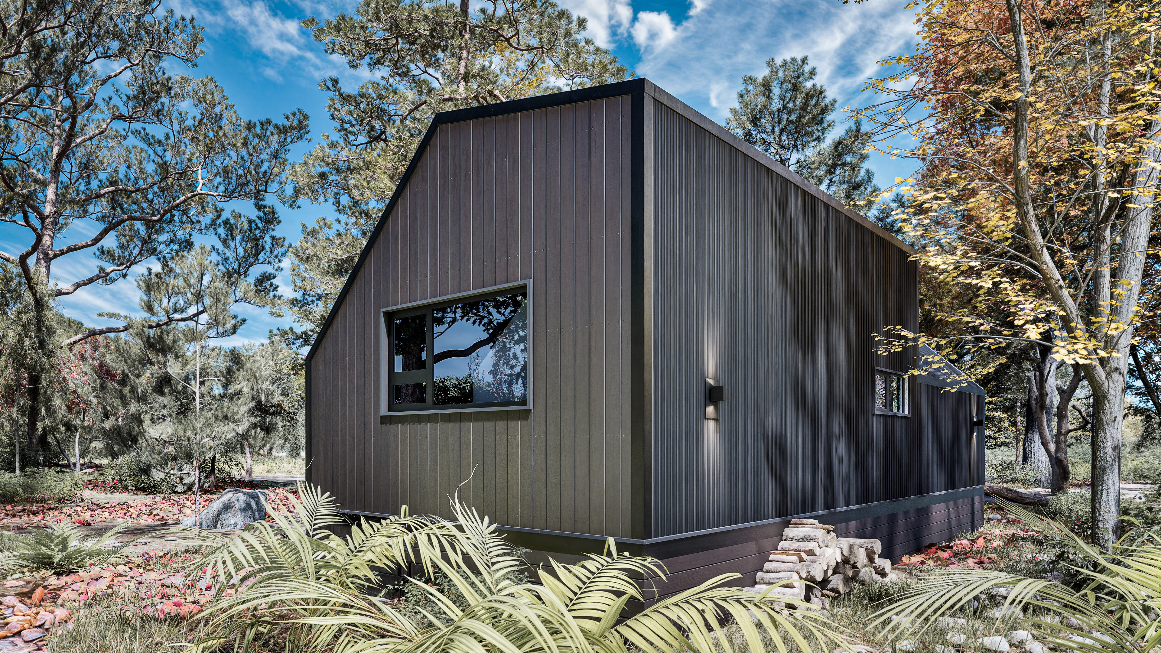 A modern dark-colored tiny house with large windows stands in a wooded area, surrounded by trees, plants, and scattered rocks under a partly cloudy sky.