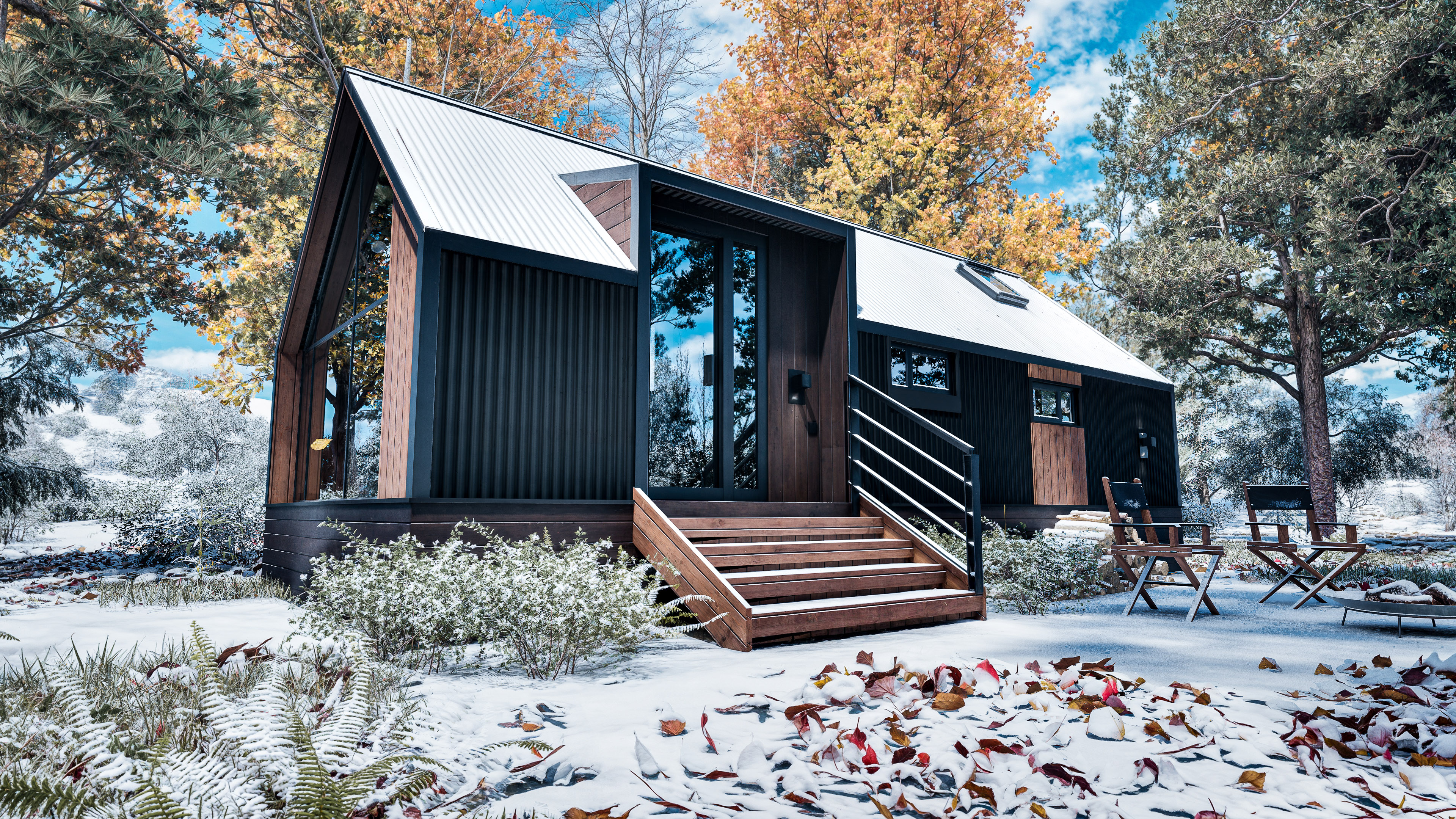 A modern tiny house with black siding and large windows sits among autumn trees and light snow, with outdoor chairs and fallen leaves visible in the yard.