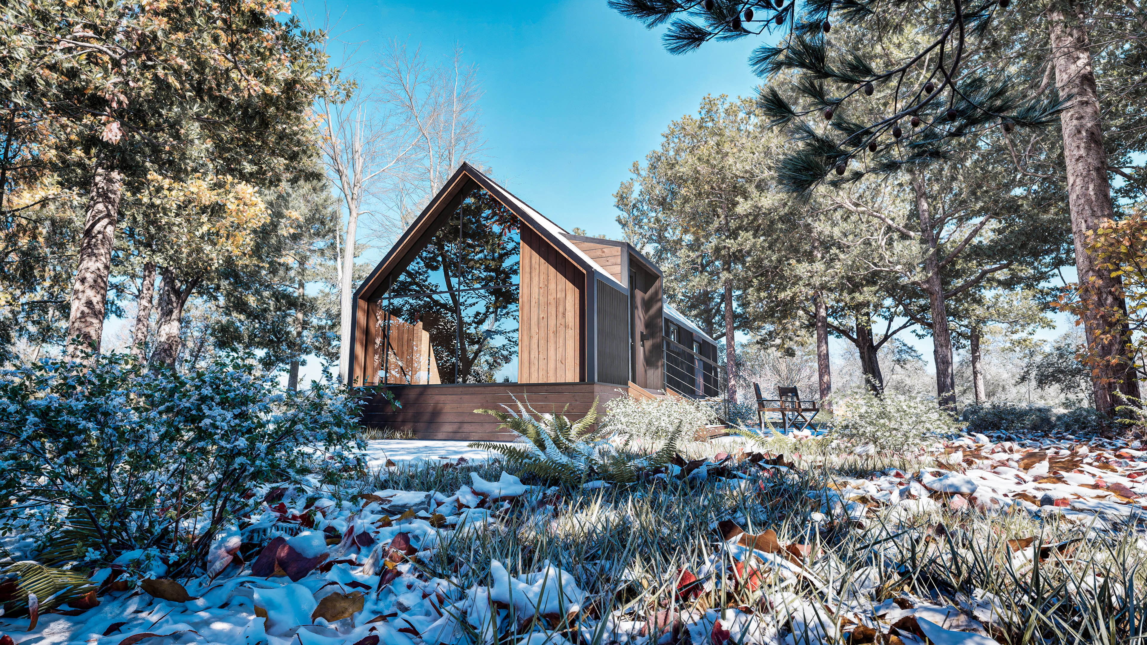 A modern wooden cabin stands among trees in a forest, with patches of snow and fallen leaves covering the ground under a clear blue sky.