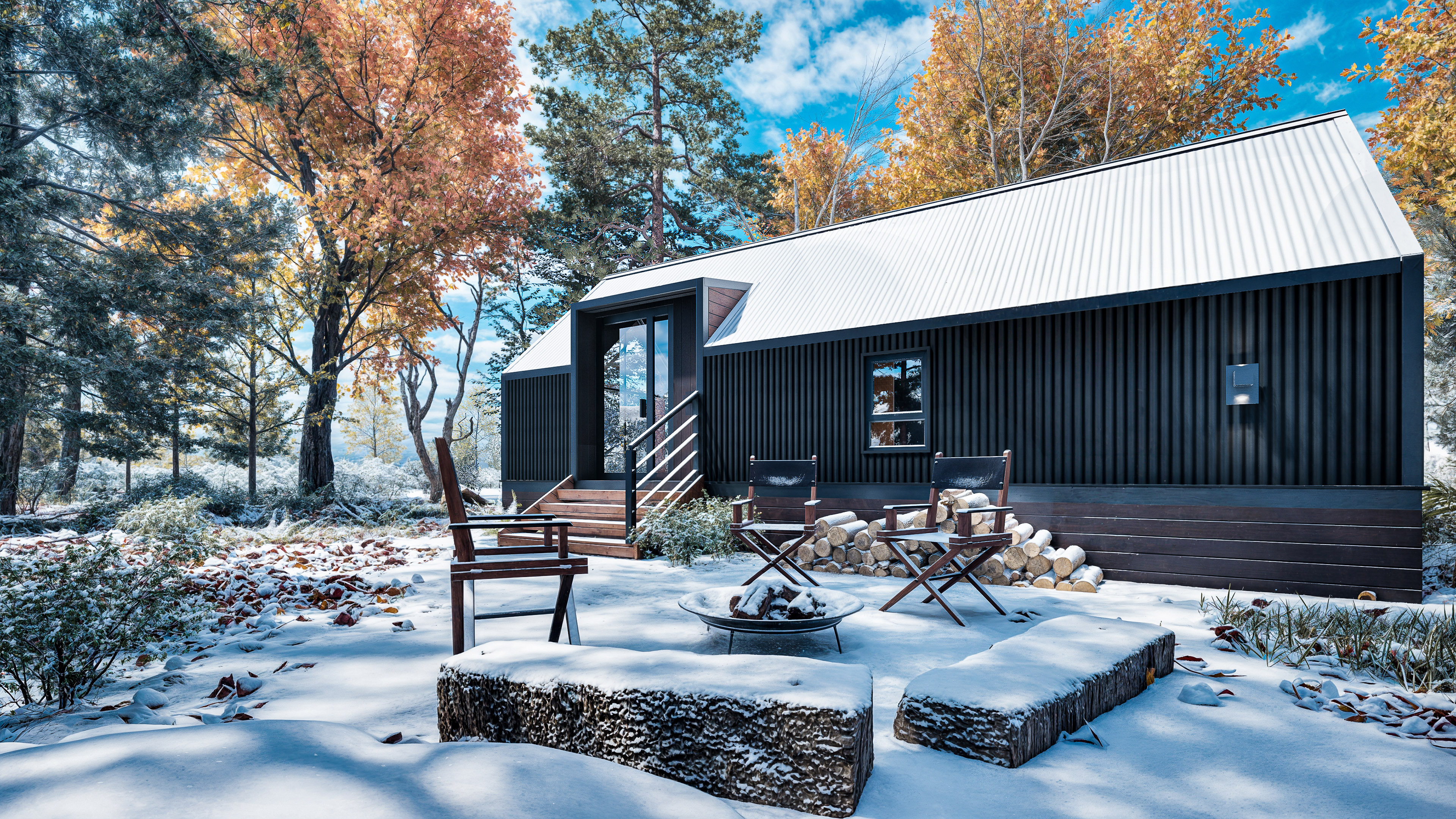 A modern black cabin with a metal roof sits in a snowy forest clearing, surrounded by autumn trees. Three chairs and a fire pit.