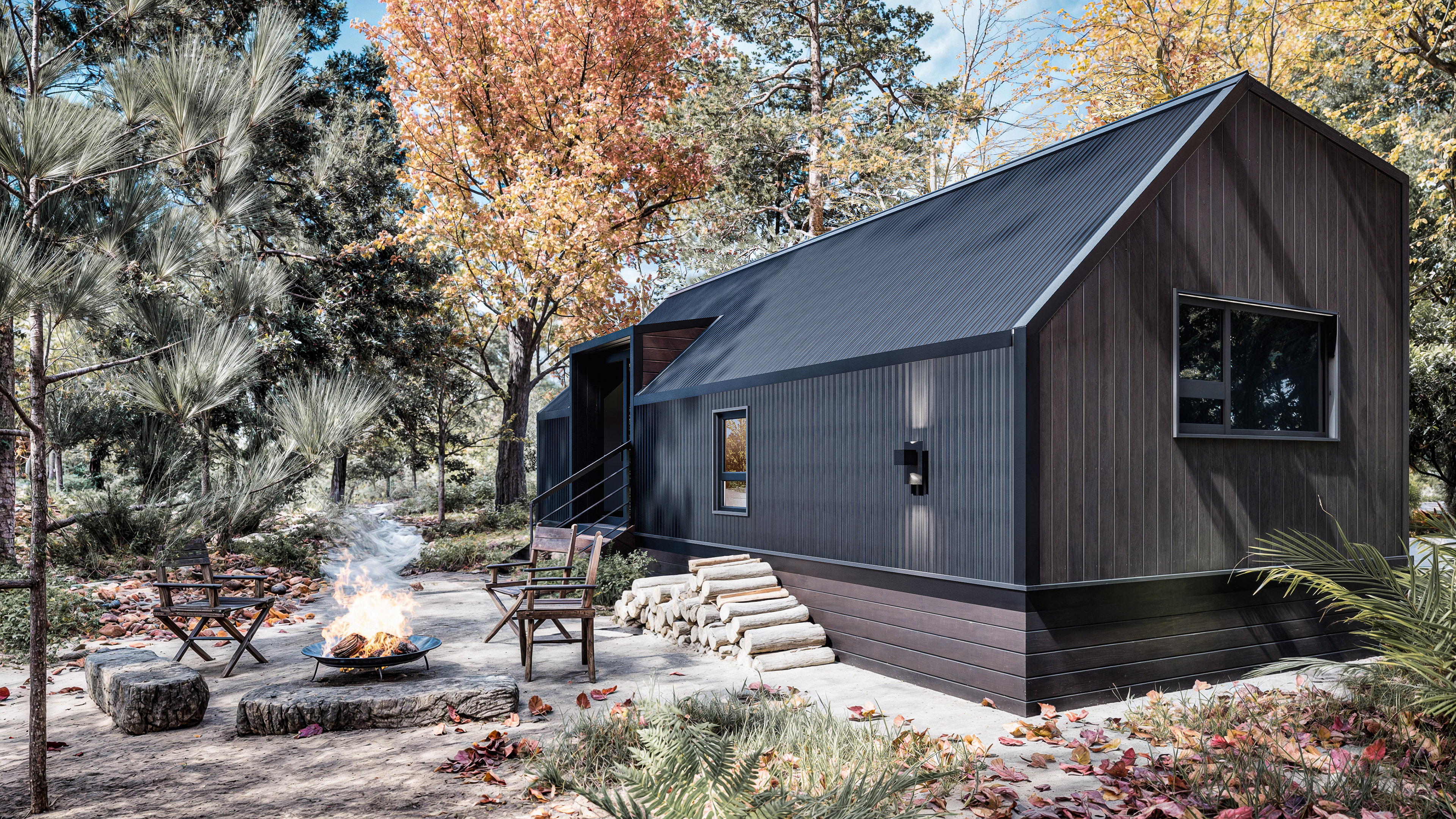 A modern dark wood cabin with a metal roof sits among autumn trees, with a fire pit and three chairs arranged on stone pavers in the yard.