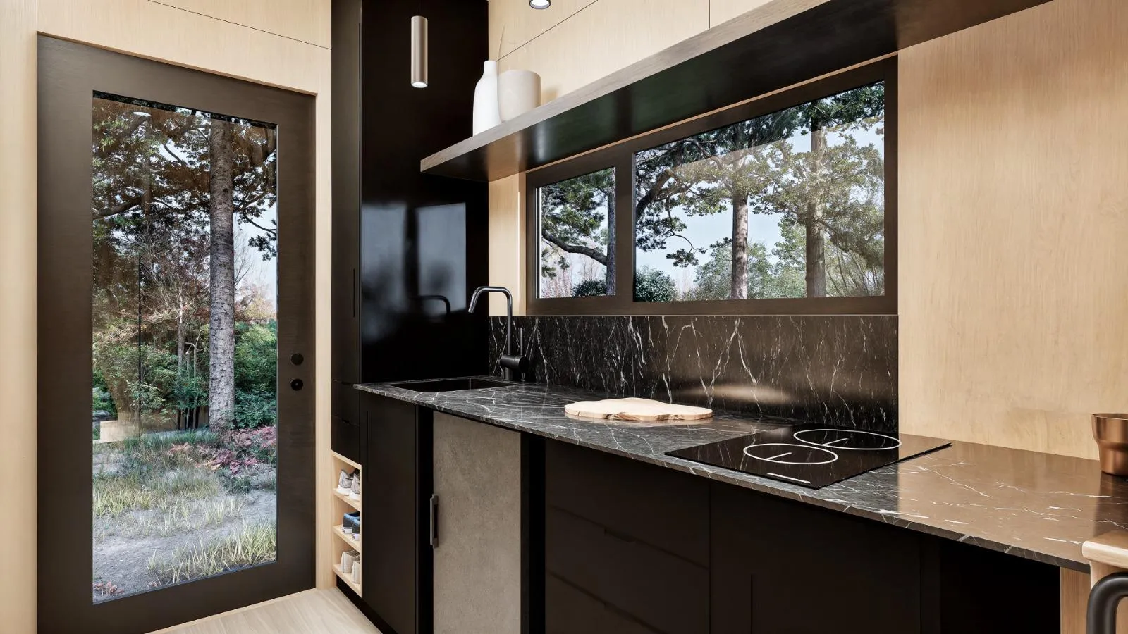 Modern cabin kitchen with black cabinets, marble countertop, induction cooktop, and sink. Large window and glass door provide views of trees and greenery outside.