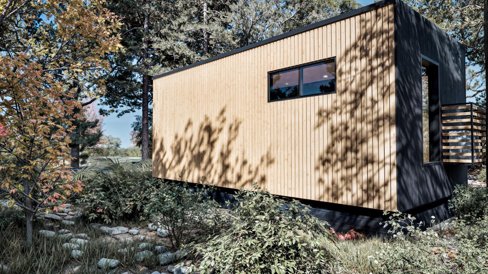 A small, modern cabin with vertical wooden siding and a single rectangular window sits among trees and plants in a natural outdoor setting.