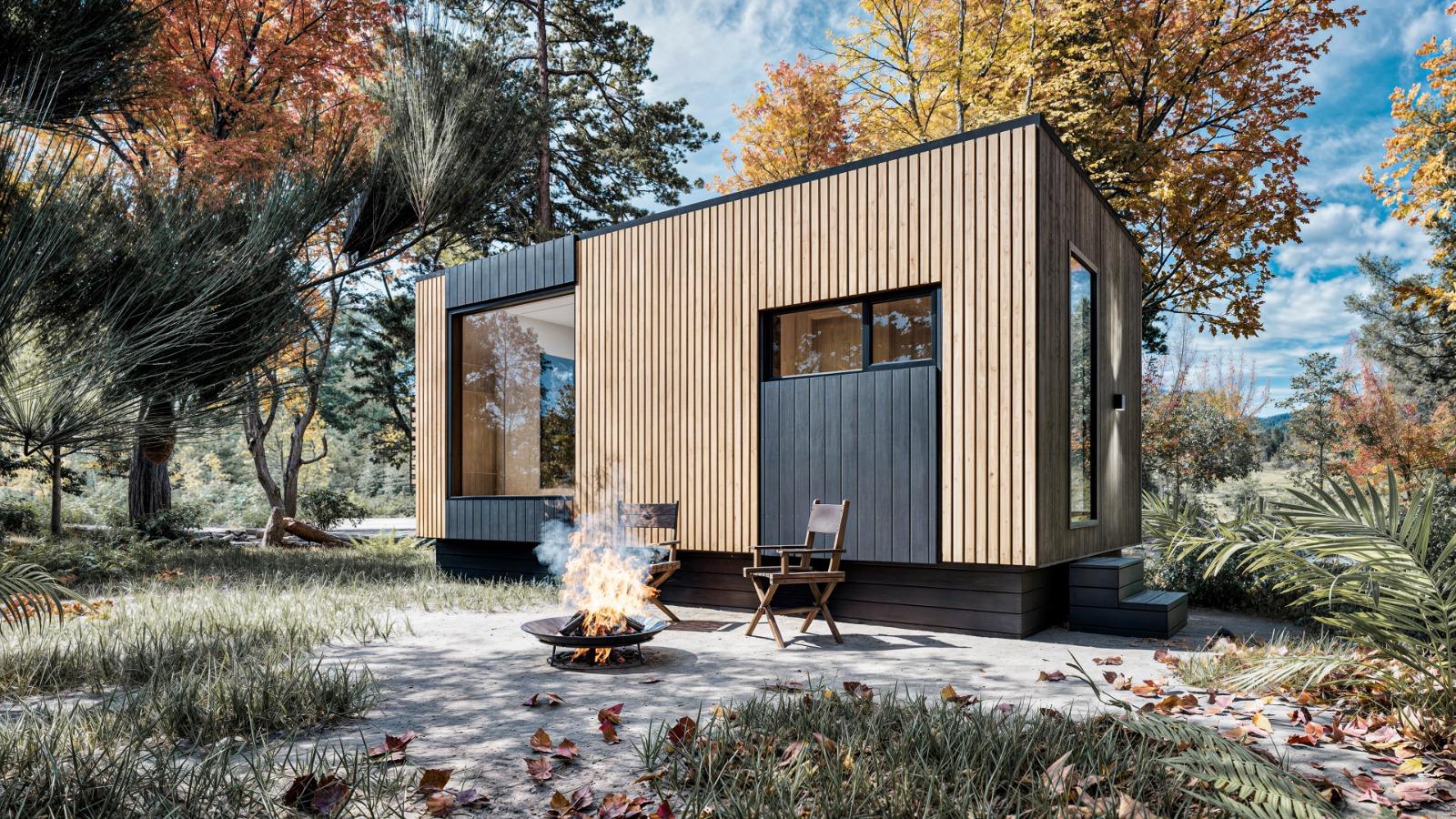 A modern cabin with wood paneling sits in a grassy yard surrounded by autumn trees, with two chairs and a fire pit in the foreground.