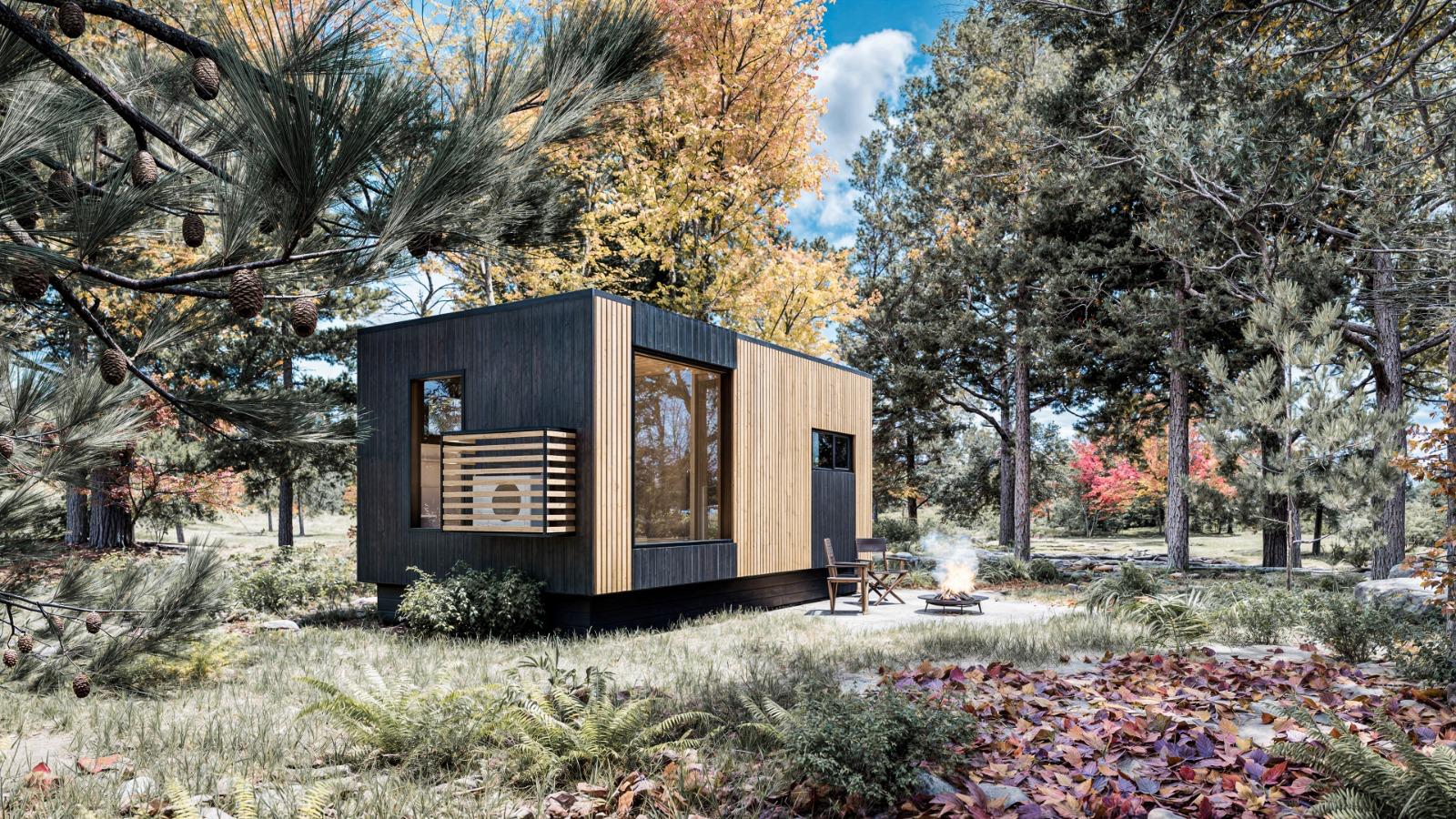 A small modern cabin with wood and dark paneling sits in a forest clearing, surrounded by trees and autumn foliage, with two chairs and a firepit outside.