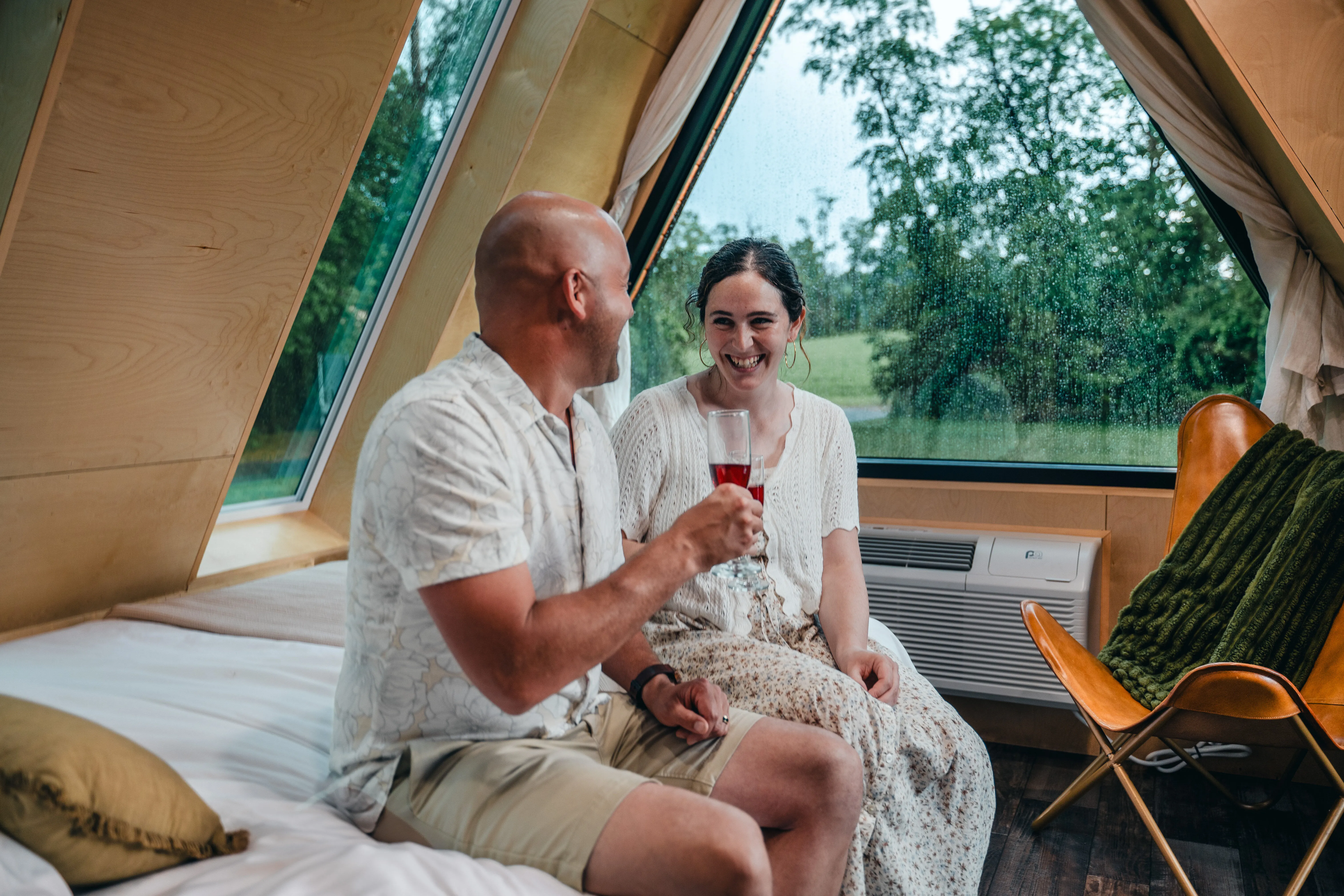 A man and woman sit on a bed in a modern cabin with large triangular windows, smiling and holding drinks while looking at each other.