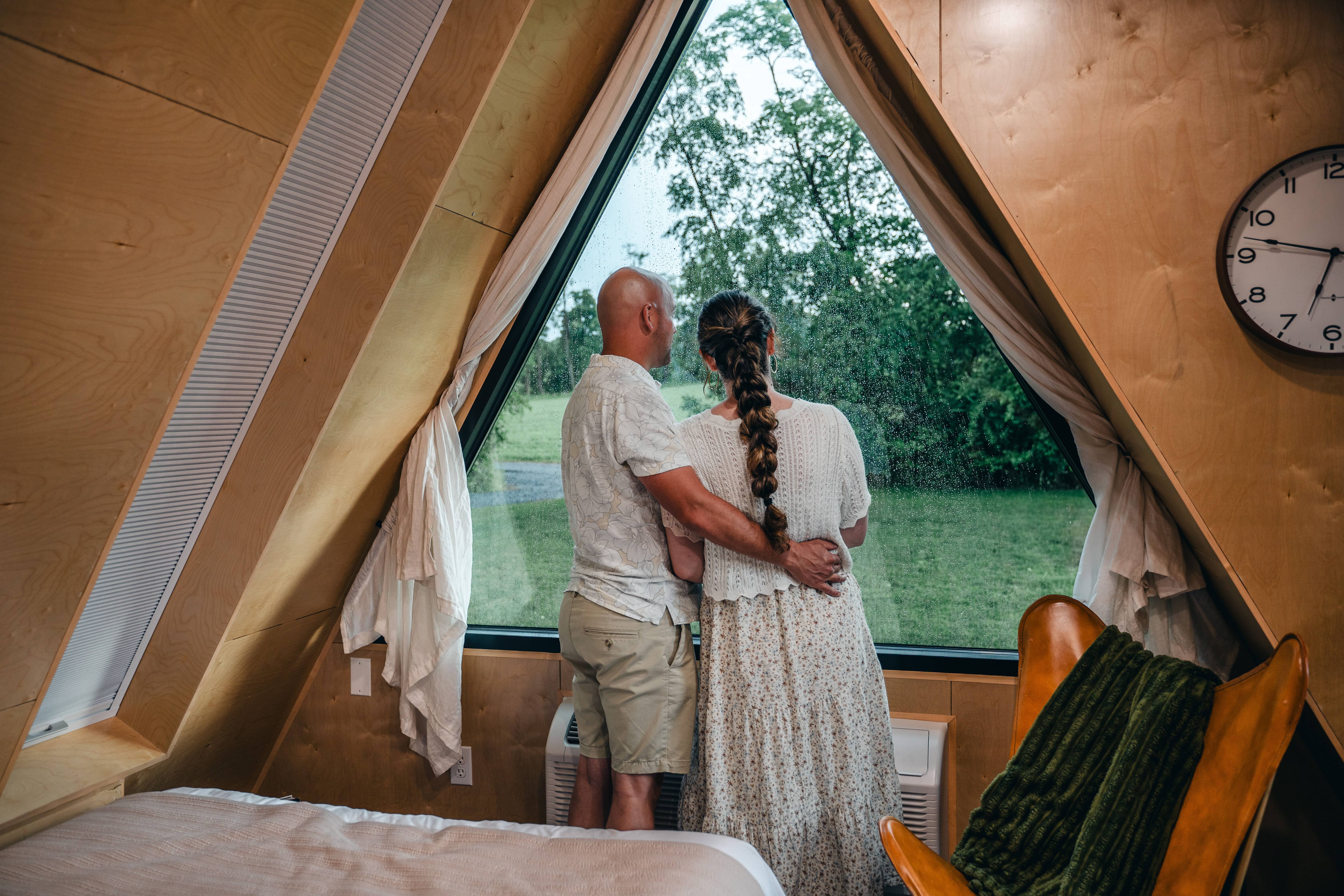 A couple stands arm in arm inside an A-frame cabin, looking out a large window at a grassy landscape on a rainy day.