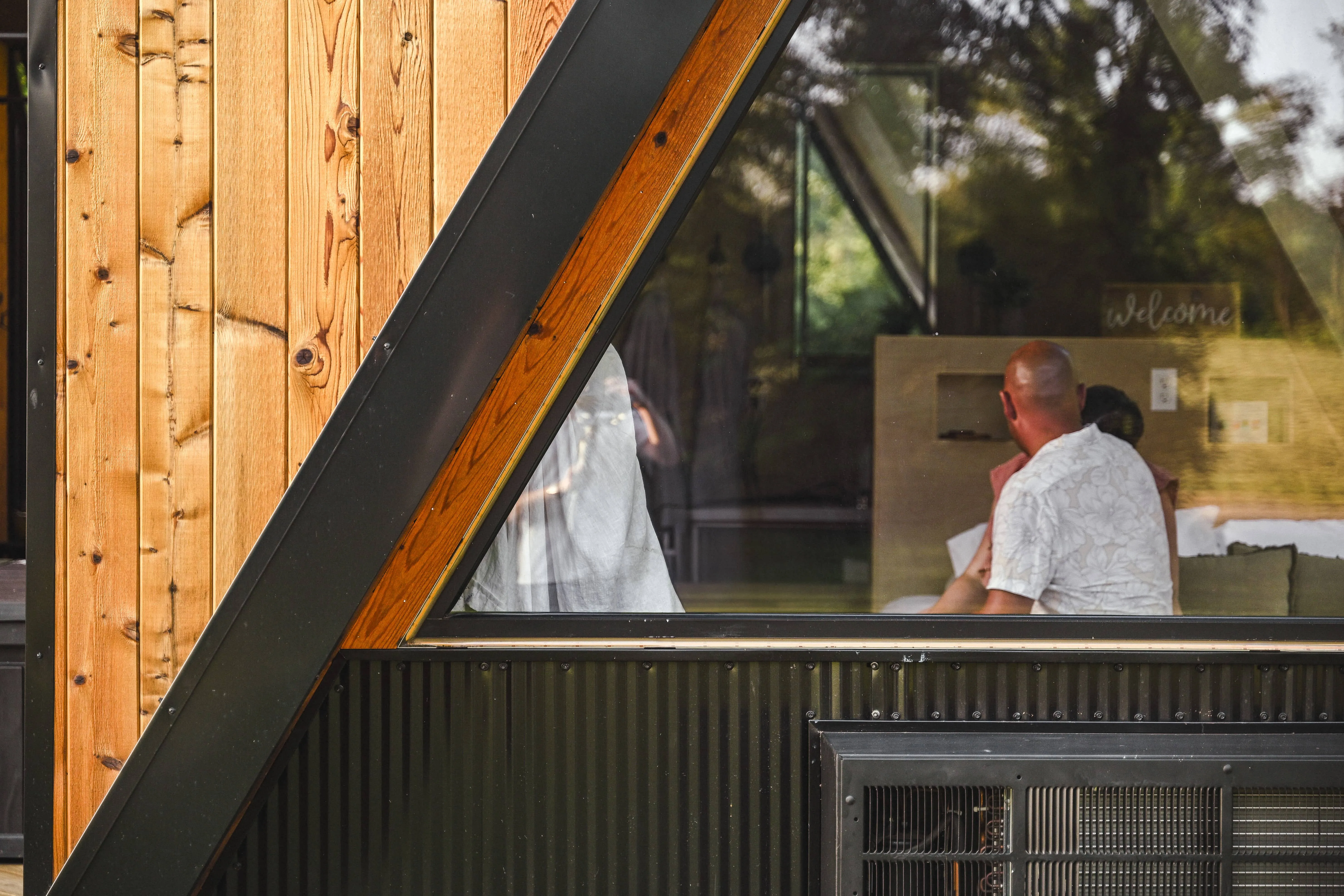 People sit inside an A-frame building with wood and metal exterior, visible through a large window reflecting outdoor greenery.