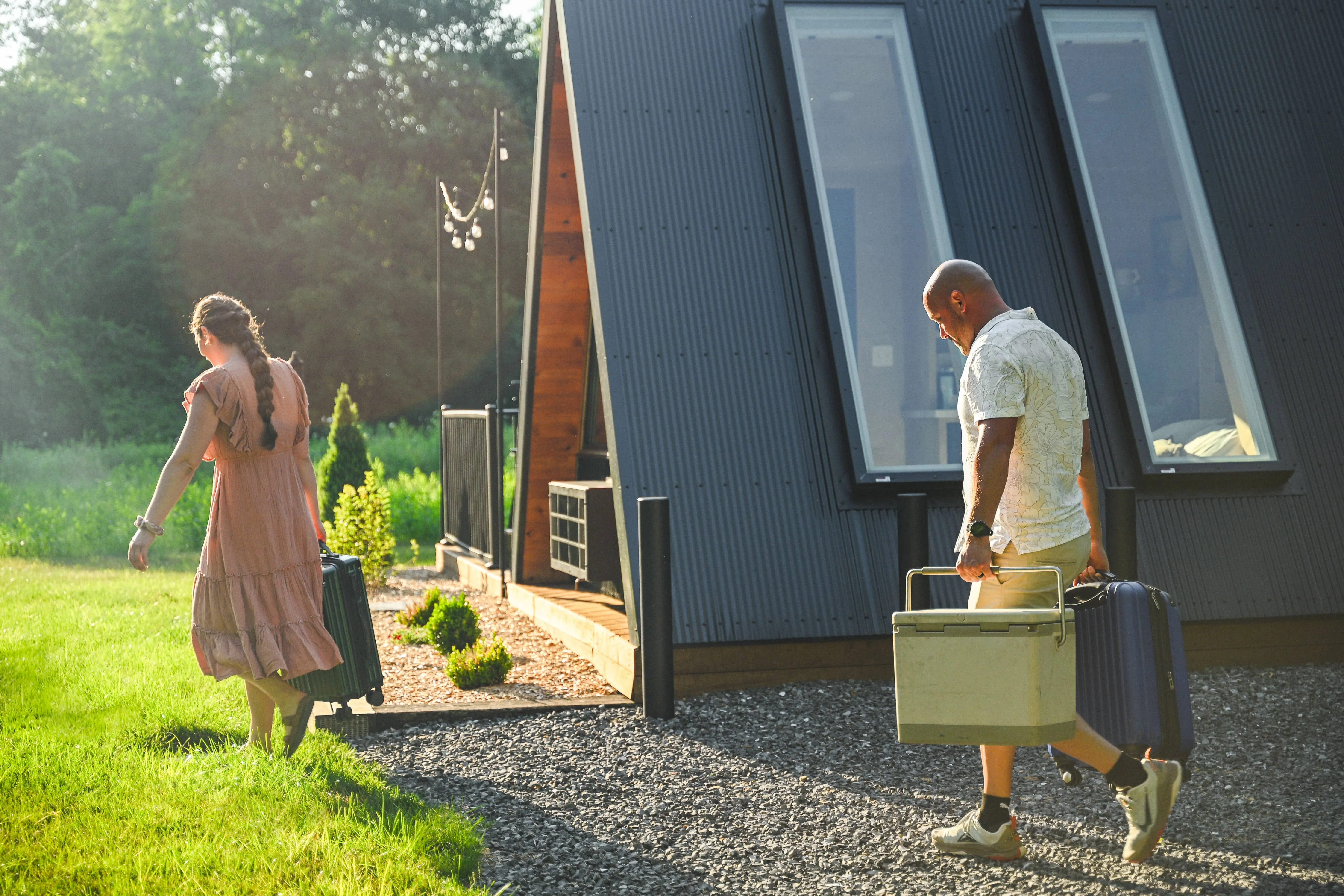 Two people carry luggage and a cooler as they walk toward a modern cabin in a grassy, sunlit outdoor setting.