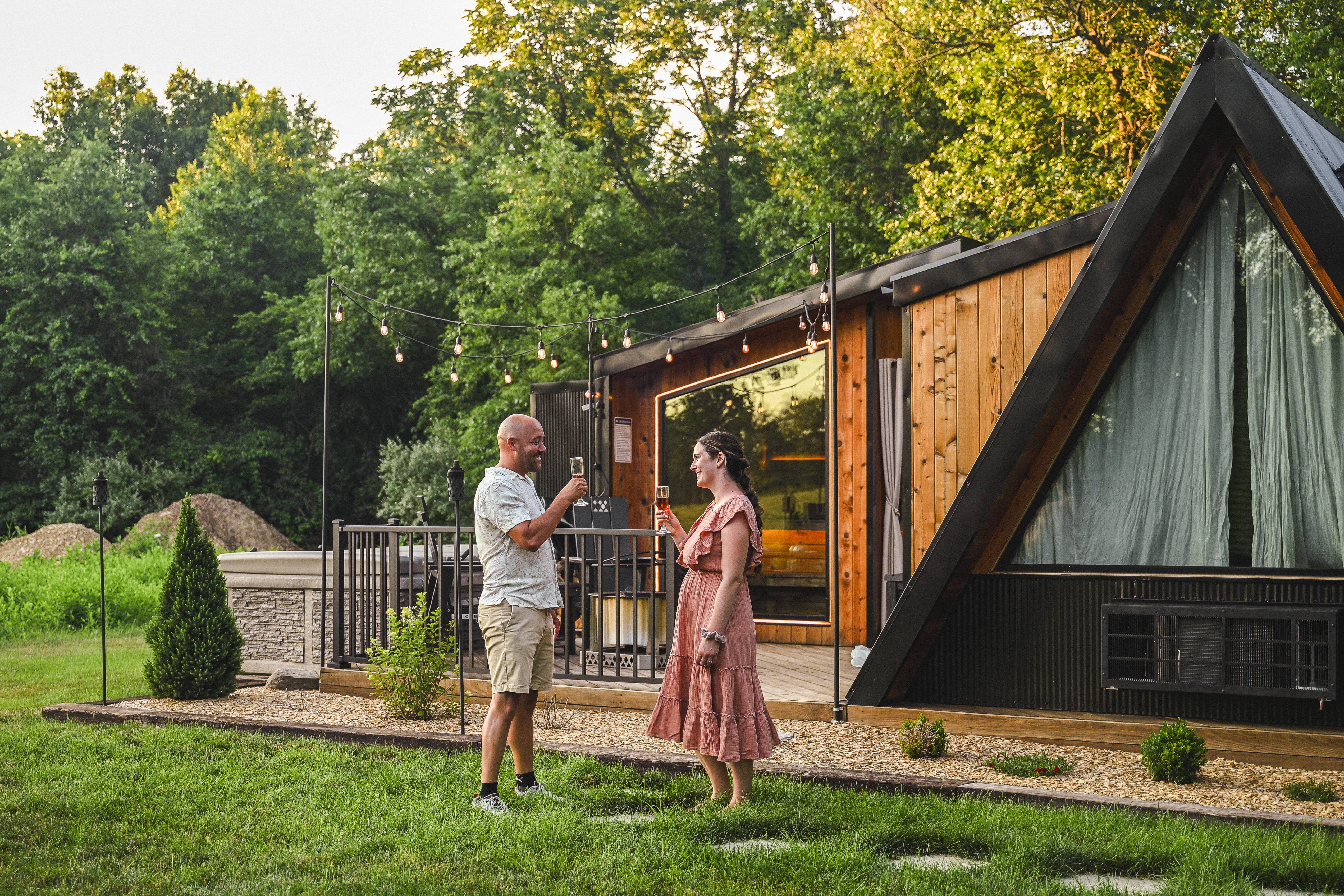 Two people stand on grass outside a modern A-frame house drinking champagne.