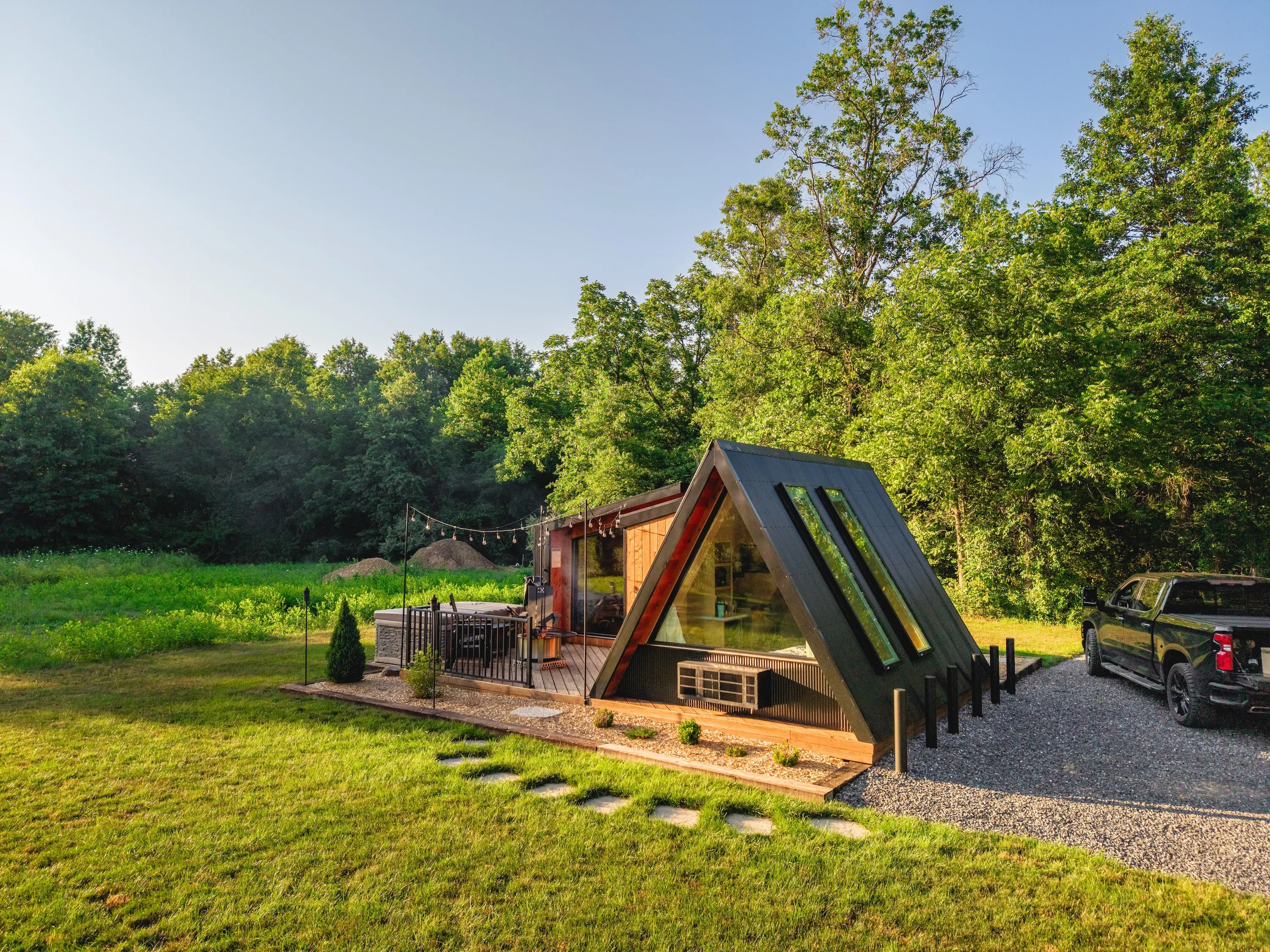 A-frame tiny house with large windows next to a patio, surrounded by grass, trees, and a parked black truck on a gravel driveway.