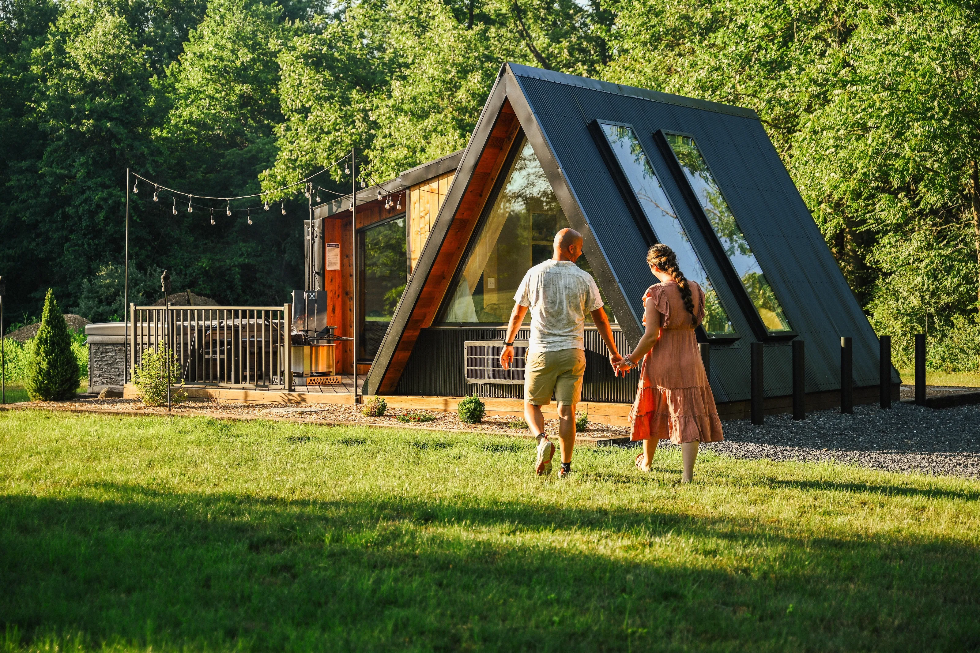 Two people hold hands while walking toward a modern A-frame tiny house with large windows, surrounded by grass, trees, and outdoor string lights.