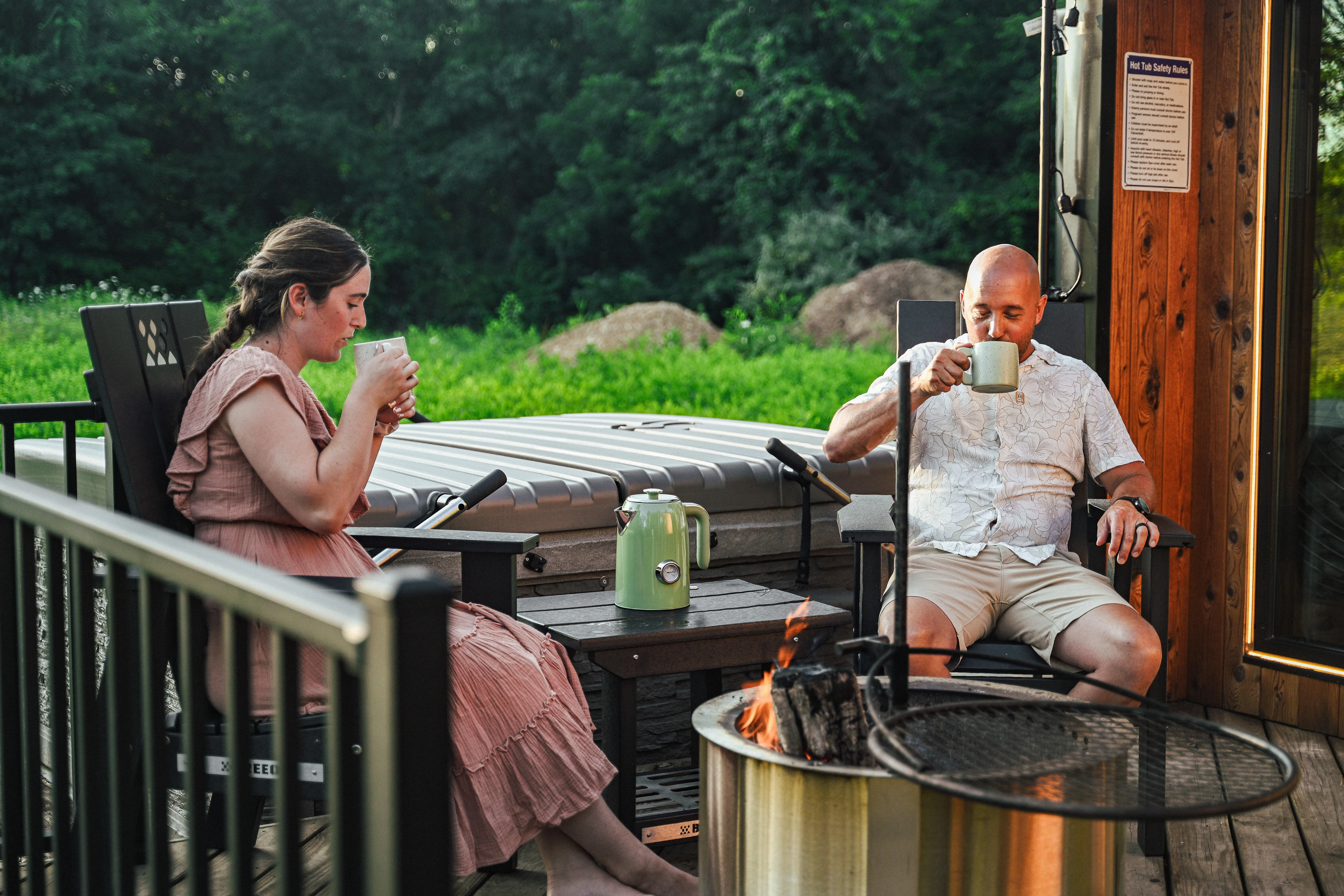 Two people sit on a porch drinking from mugs near a fire pit, with a green field and trees in the background.