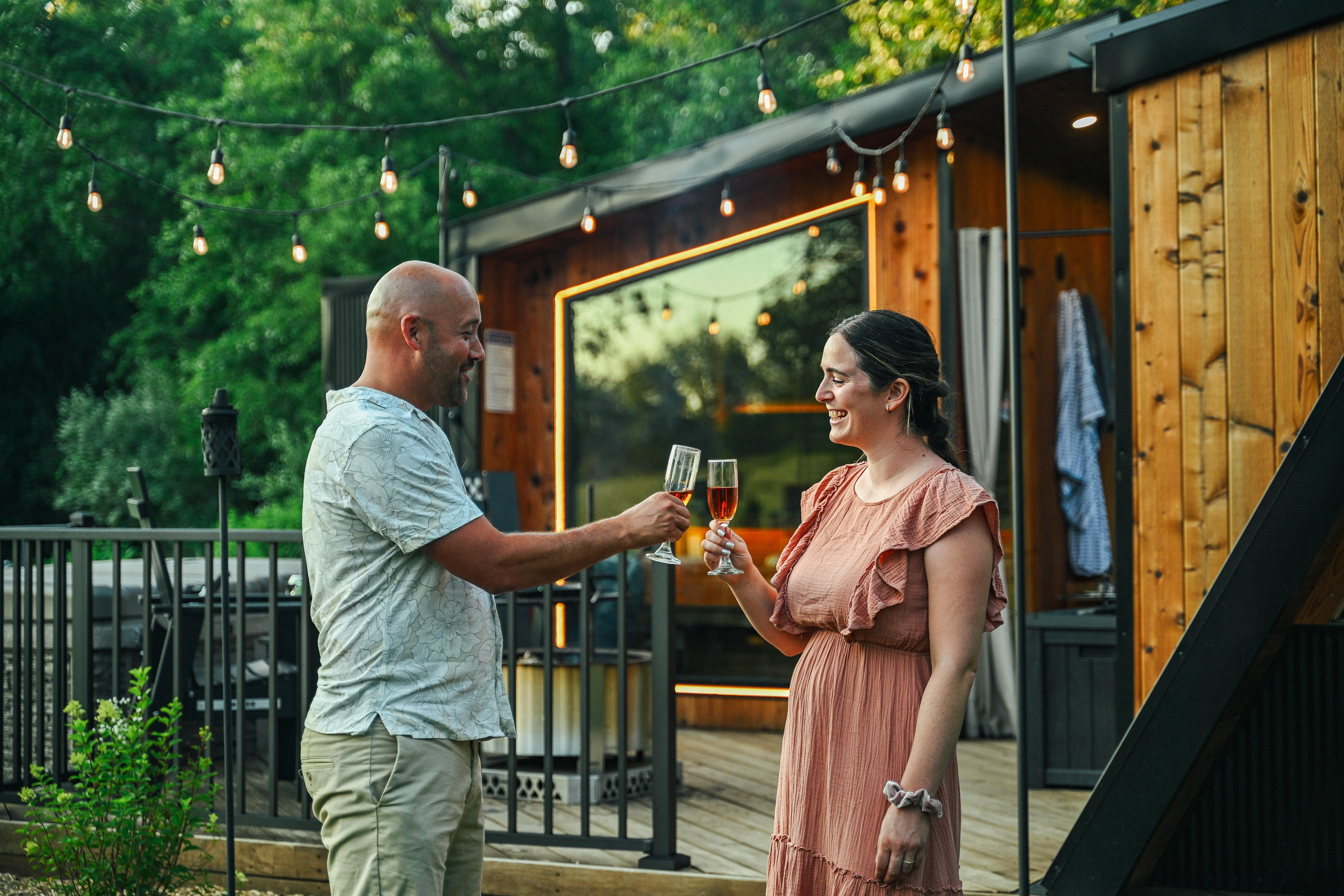 A man and woman clink glasses while standing outside a modern wooden cabin under string lights on a sunny day.