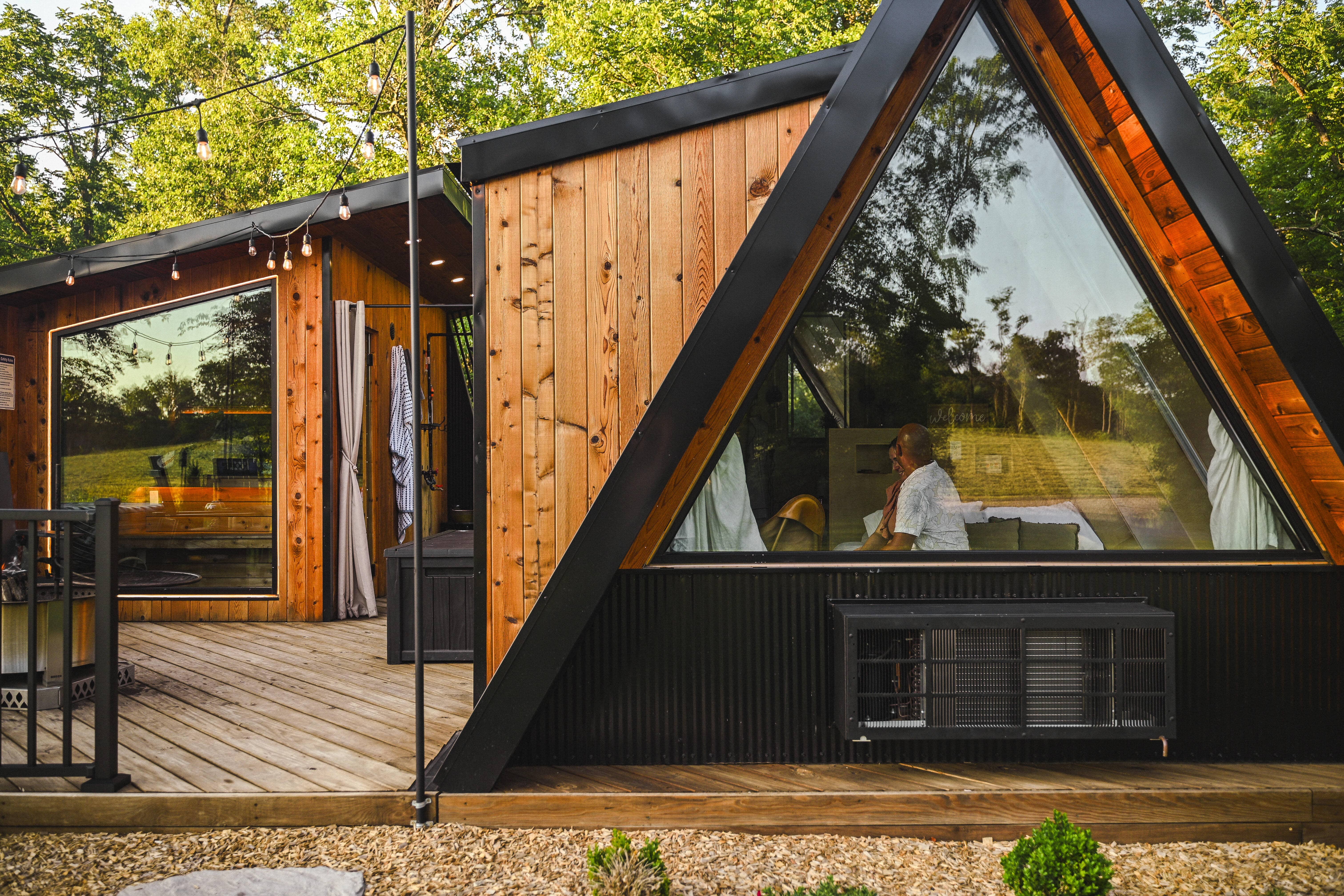 A person sits inside a modern A-frame cabin with large triangular windows, surrounded by trees and outdoor lights, with a wooden deck in the foreground.