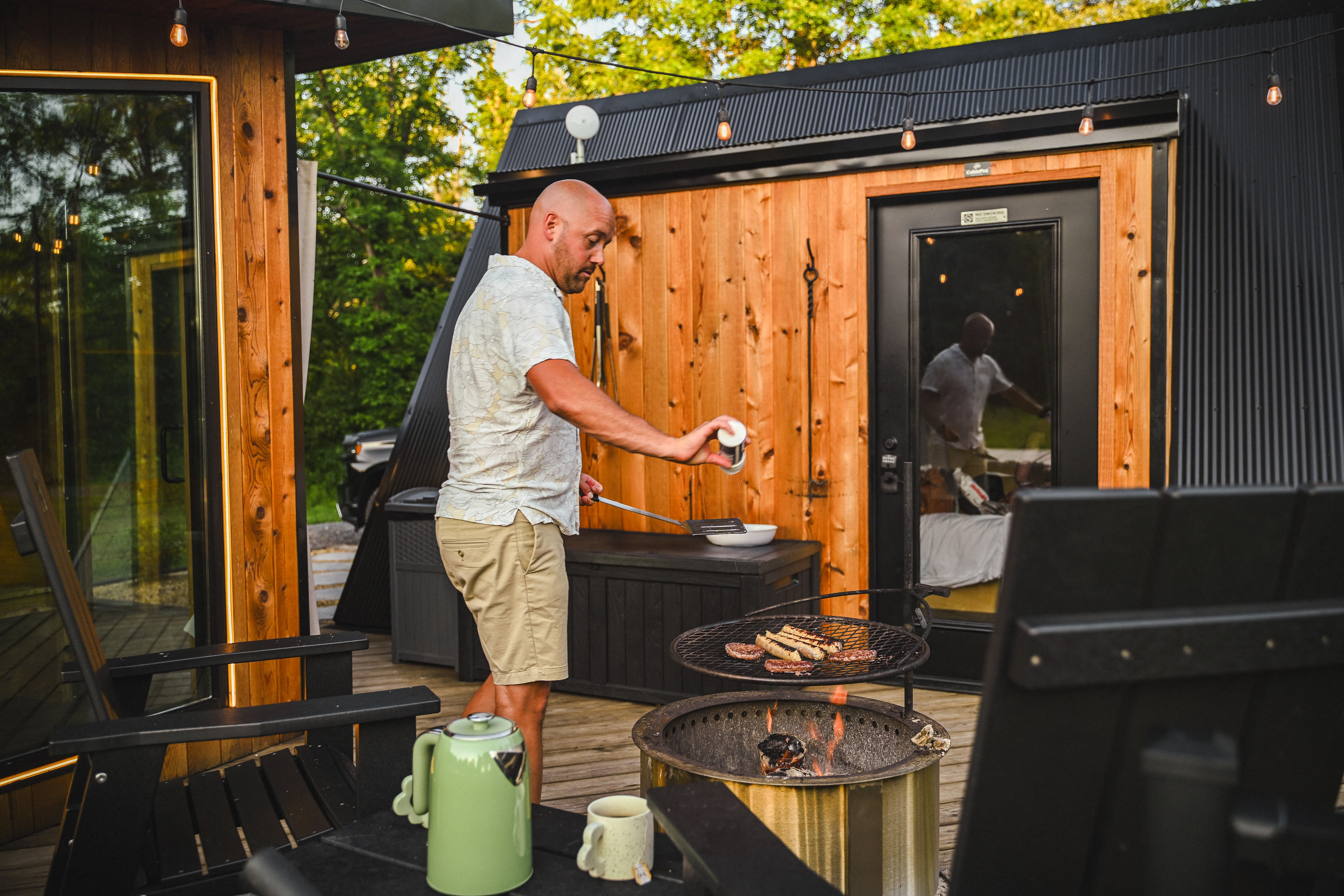 A man grills sausages on an outdoor barbecue next to a modern wooden cabin. A green kettle and cups are on the table nearby.