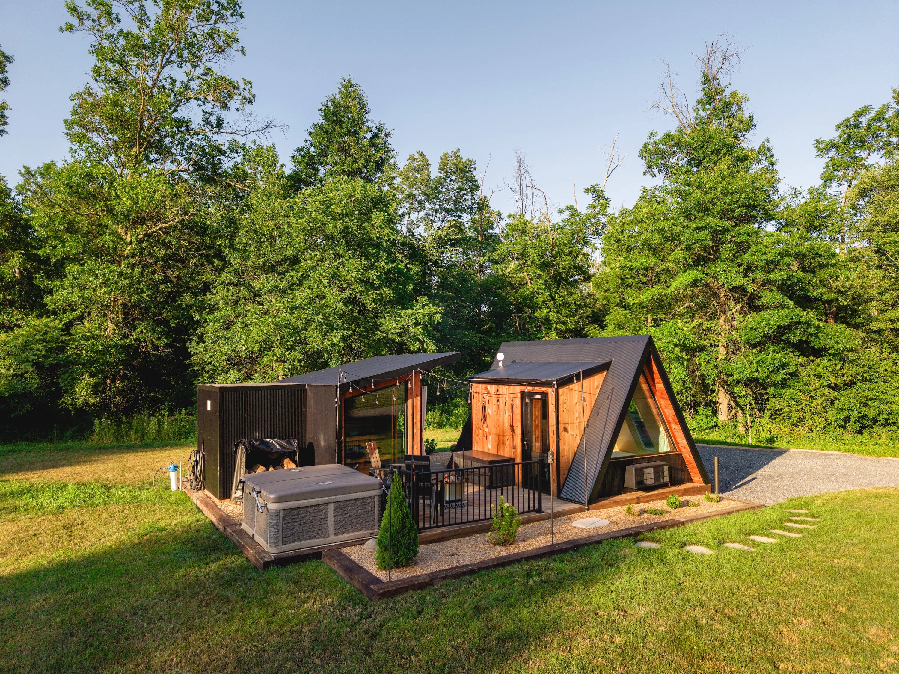 A-frame cabin with large windows, black accents, patio, hot tub, and outdoor seating, surrounded by grass and dense green trees under a clear blue sky.