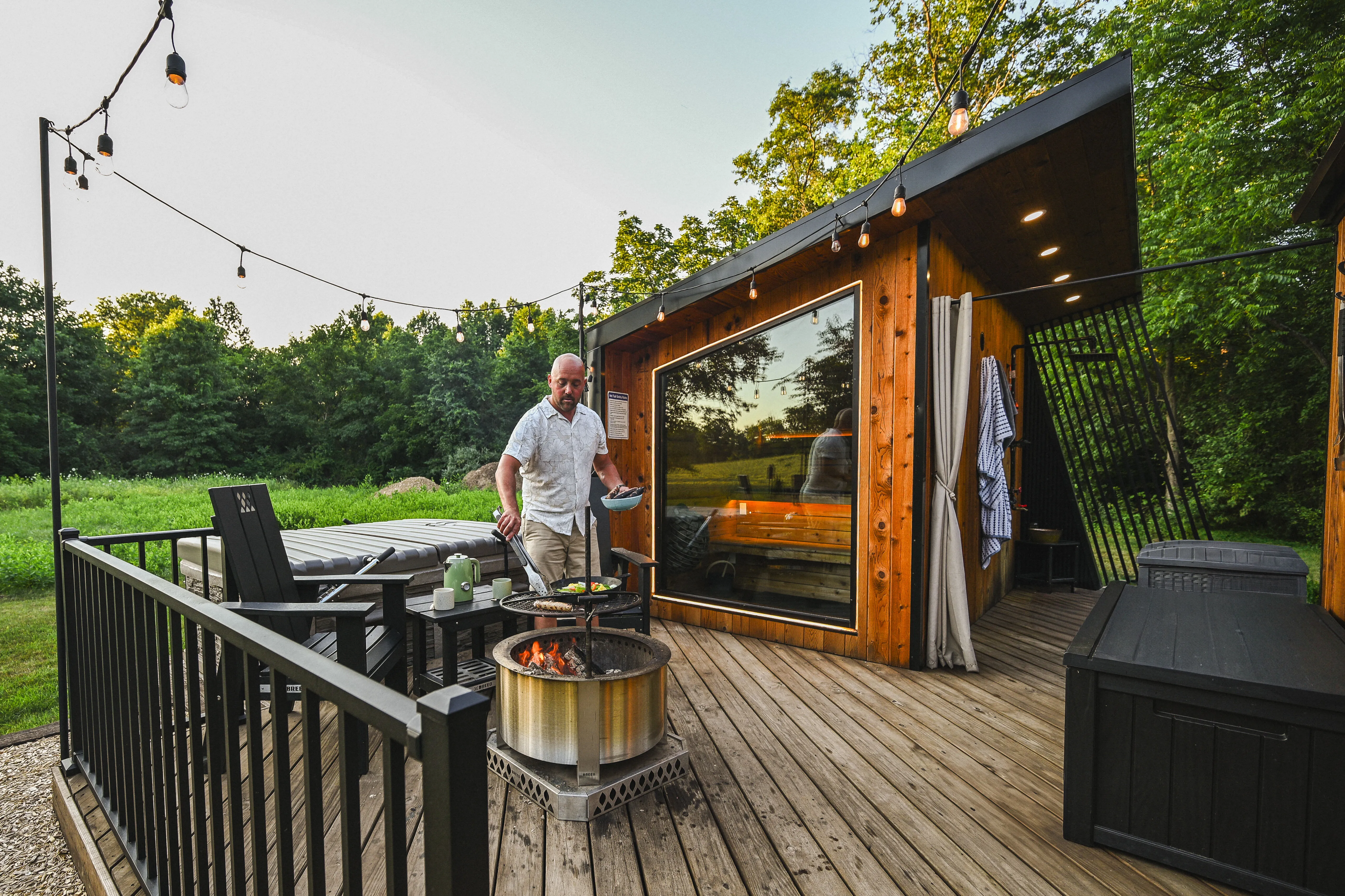 A man stands on a wooden deck next to a fire pit, outside a modern cabin surrounded by green trees and outdoor lights.