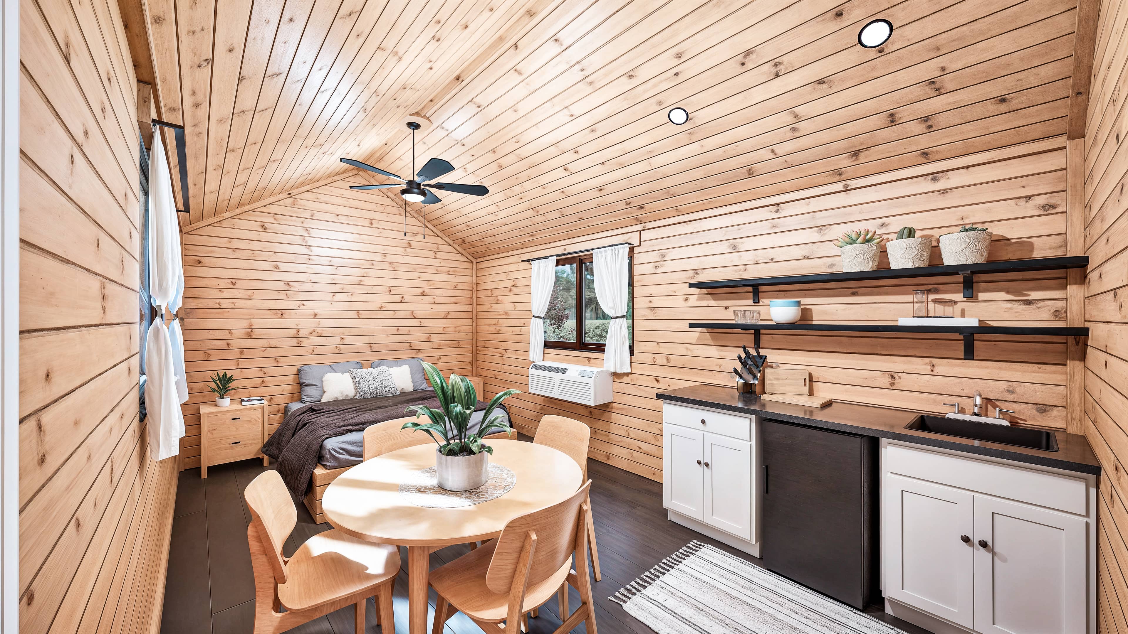 Modern wood-paneled cabin interior with a bed, round dining table, kitchenette, open shelves, plants, and a ceiling fan.