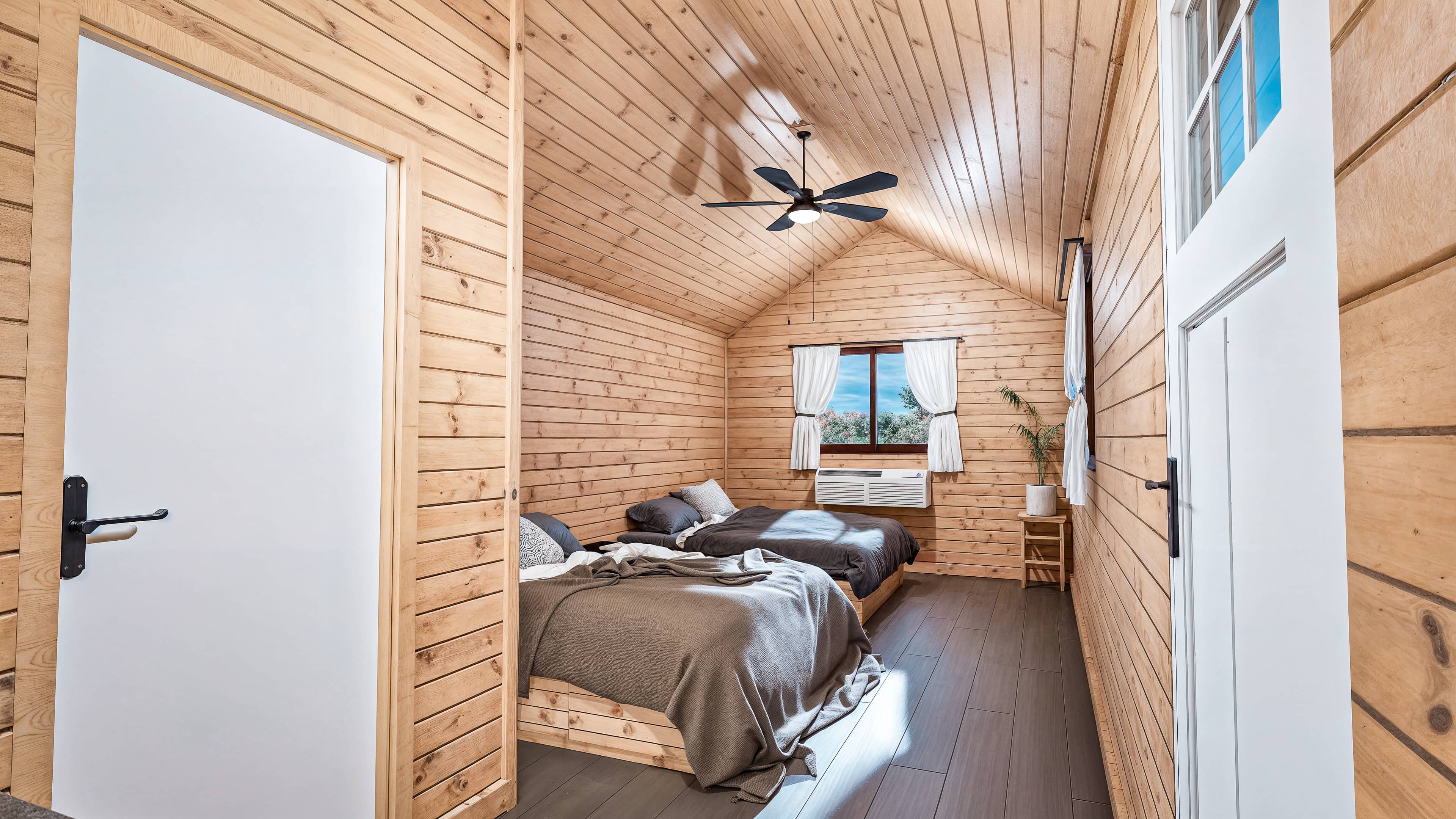 A wood-paneled bedroom with two beds, gray bedding, a ceiling fan, a window with white curtains, and a small bedside table with a plant.